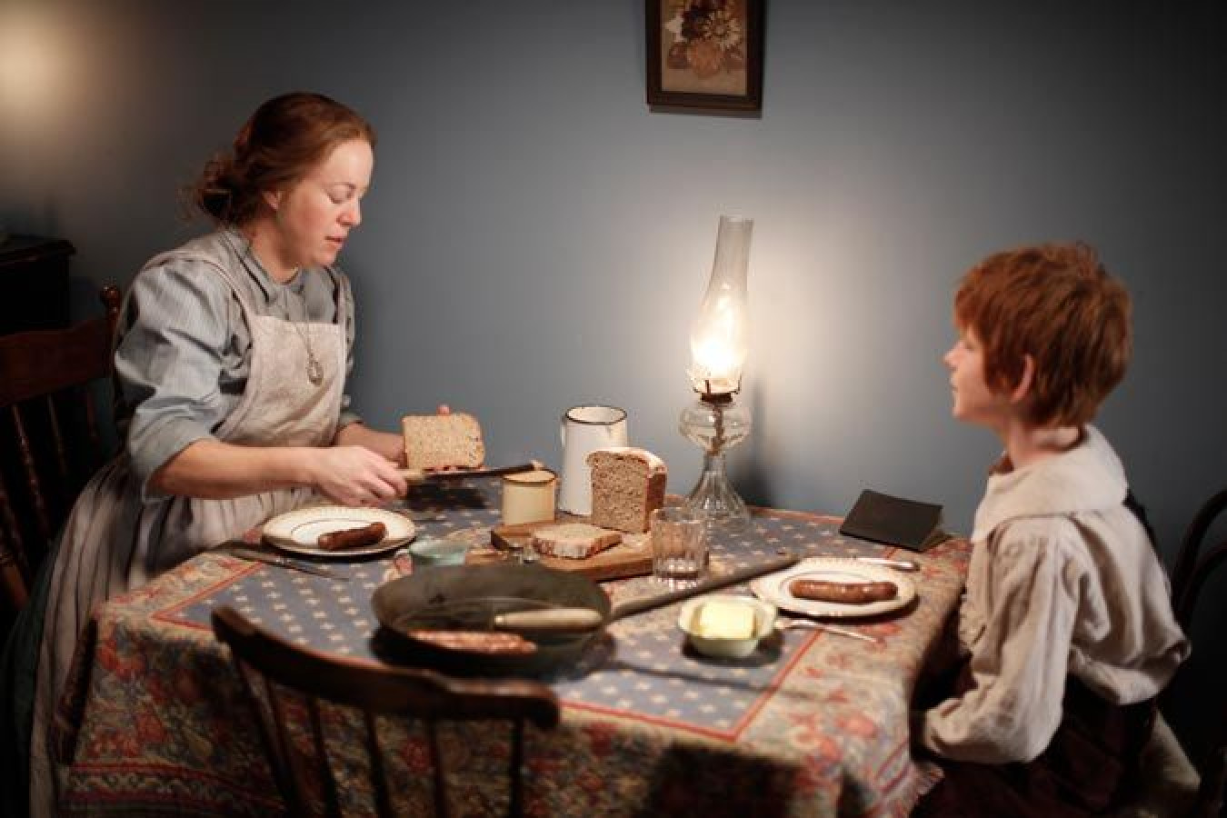 A woman in period attire carefully prepares a meal at a rustic table, spreading food on slices of bread with a child seated nearby. An oil lamp casts a warm glow in the dimly lit room, creating a cozy, historical atmosphere.