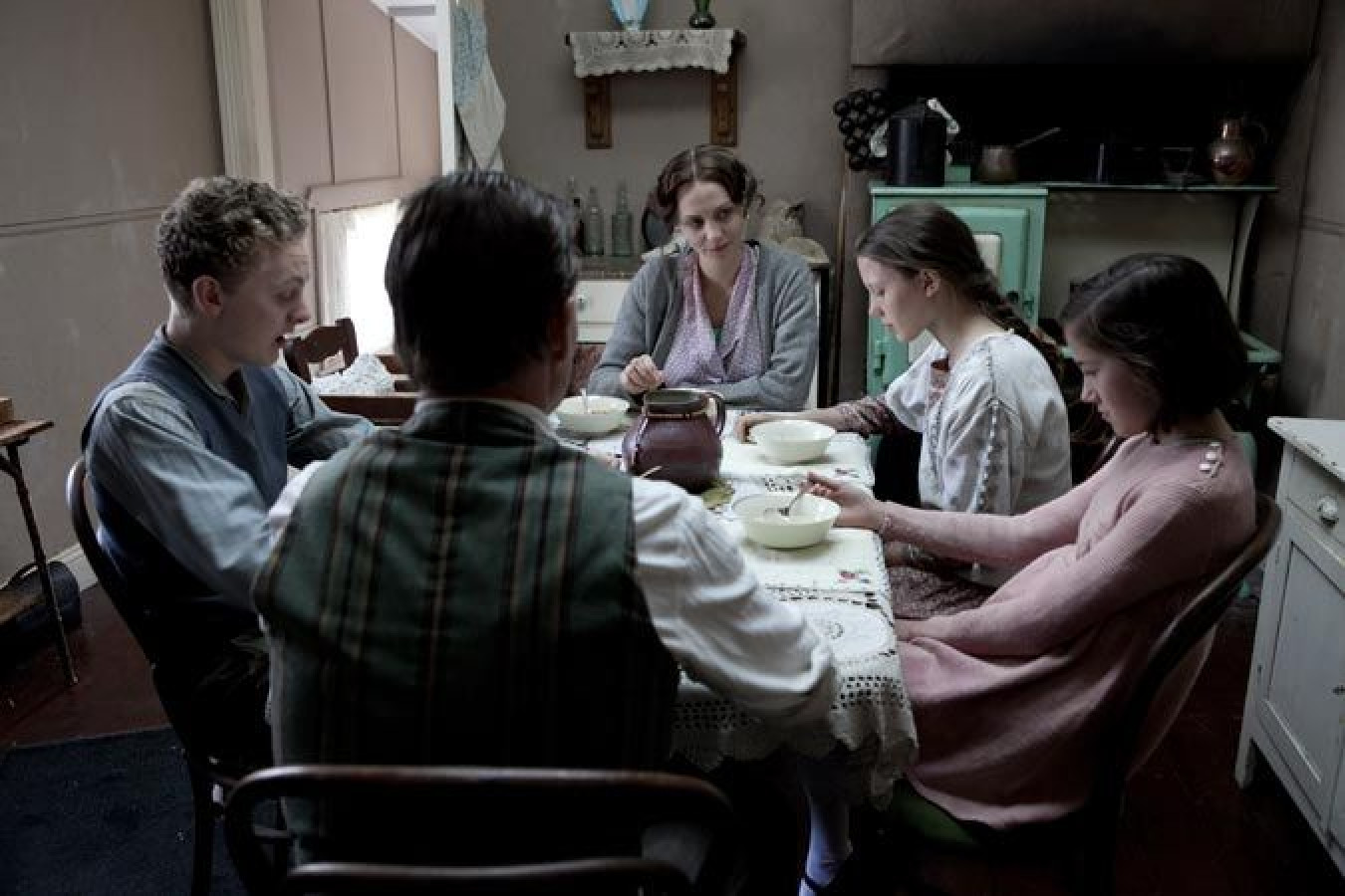 A family sits around a dining table in a modest home, sharing a meal. The scene captures four children and one adult, all engaged in their food with expressions of contemplation, surrounded by simple kitchen decor and a green stove in the background.