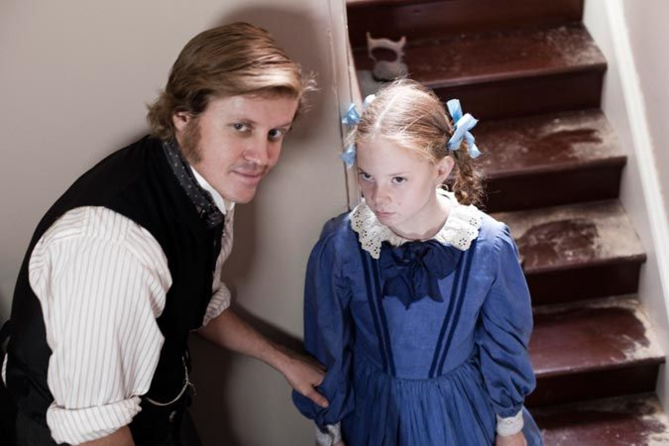 A young girl in a blue dress stands on a staircase, looking displeased while clutching a man's hand. The man, dressed in period clothing with sideburns, gazes at her, creating a tense atmosphere. Dust covers the stairs, suggesting neglect.