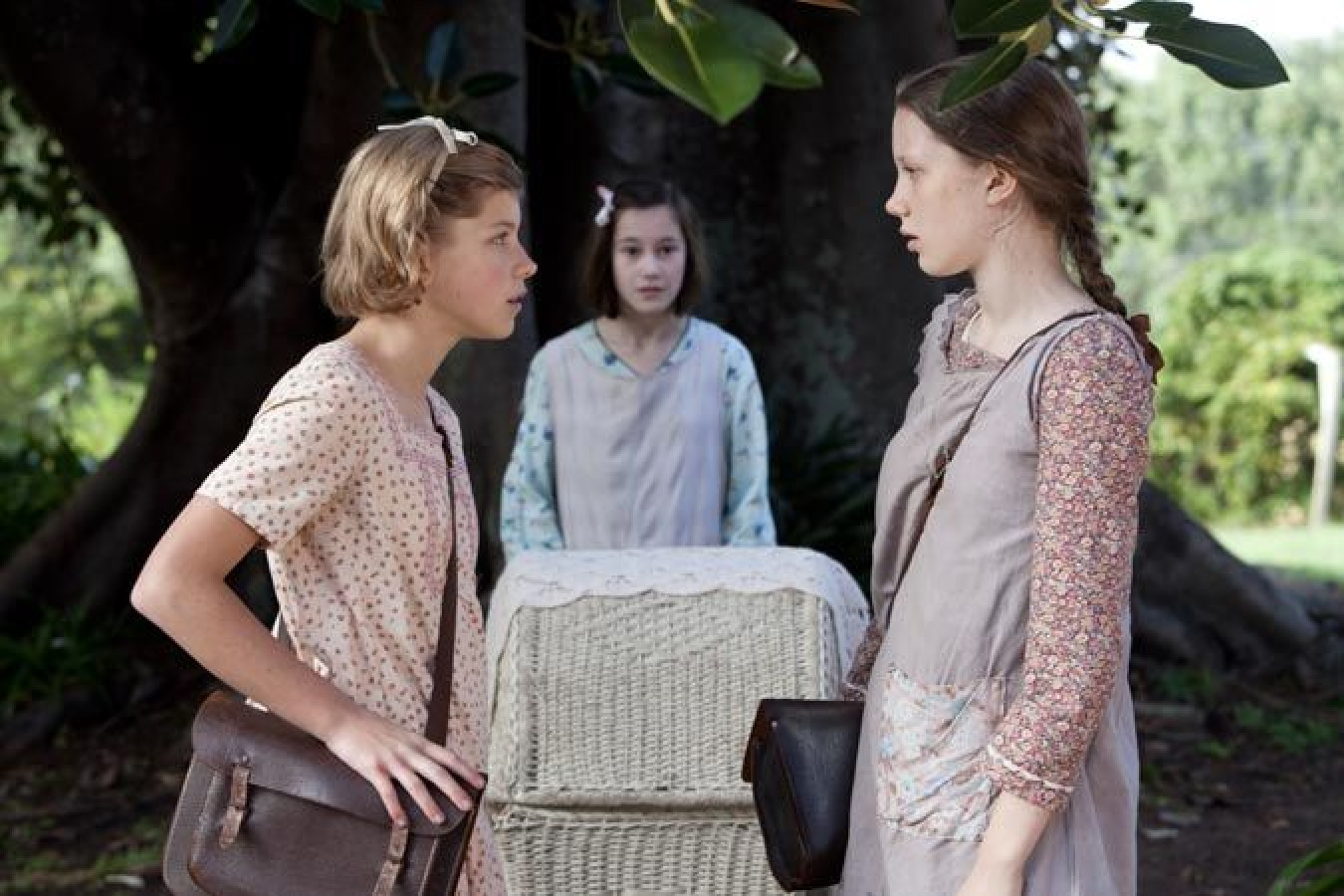 Two girls in vintage dresses stand facing each other with serious expressions, one with a satchel. A third girl in a lighter dress stands behind them, watching the confrontation. They are near a large tree, with a woven basket resting on the ground between them.