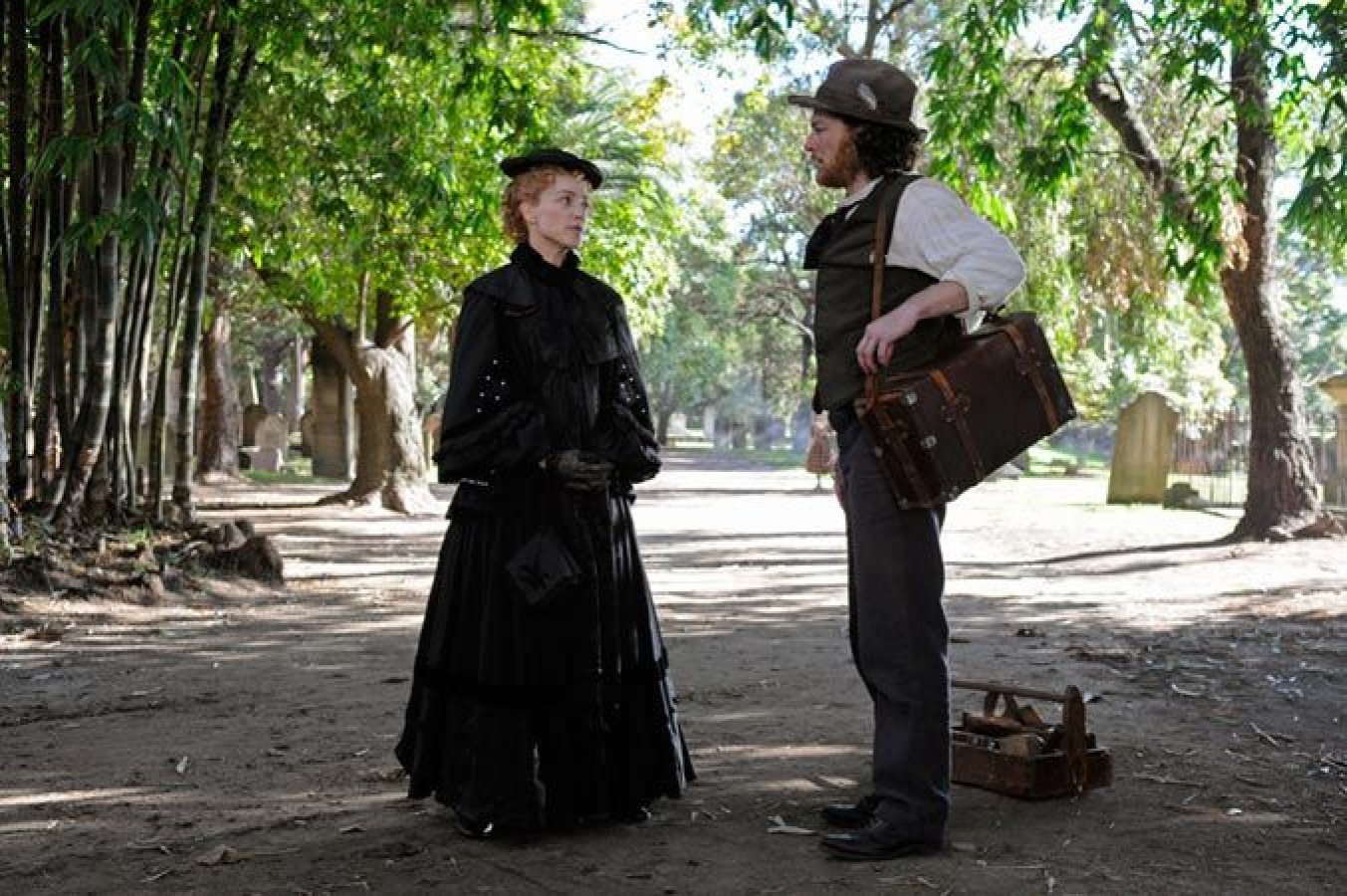 A woman dressed in a long black Victorian-style dress and hat stands in conversation with a man wearing a white shirt, vest, and a grey hat. The man holds a suitcase and is standing on a dirt path surrounded by trees and gravestones.