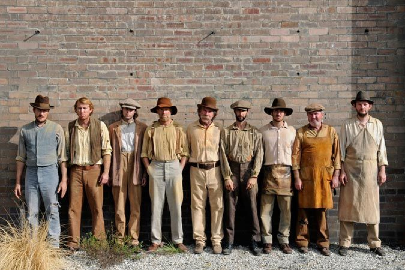 A group of nine men stands in a straight line against a brick wall, wearing light-colored clothing and wide-brimmed hats. Their expressions vary, and they seem to be dressed in a rustic style, reflecting an earlier time period.
