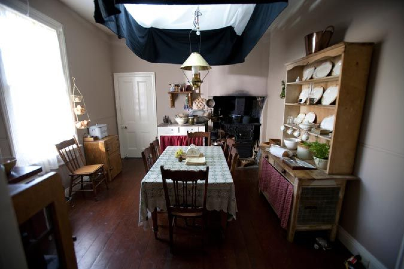 A cozy dining area features a wooden table with a lace tablecloth, surrounded by chairs. Shelves display dishware and potted plants, while a vintage lamp hangs from the ceiling. Light streams in from a window, and a rustic fireplace is visible, adding to the warm ambiance.
