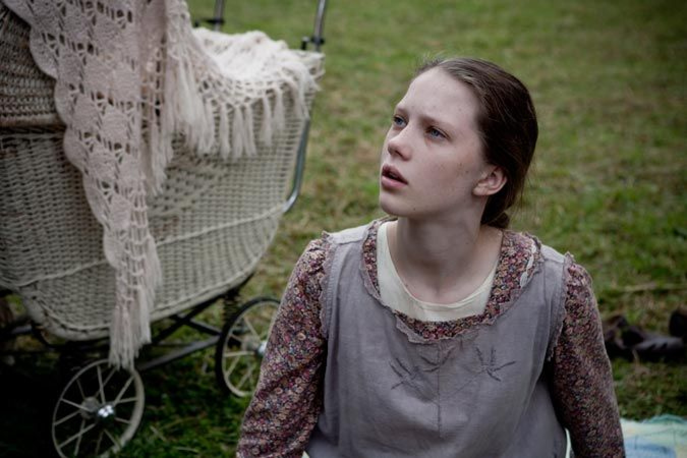 A young girl sits on the grass, looking up with a concerned expression. Behind her is an old-fashioned baby stroller draped with a knitted blanket, set against a blurred outdoor background.