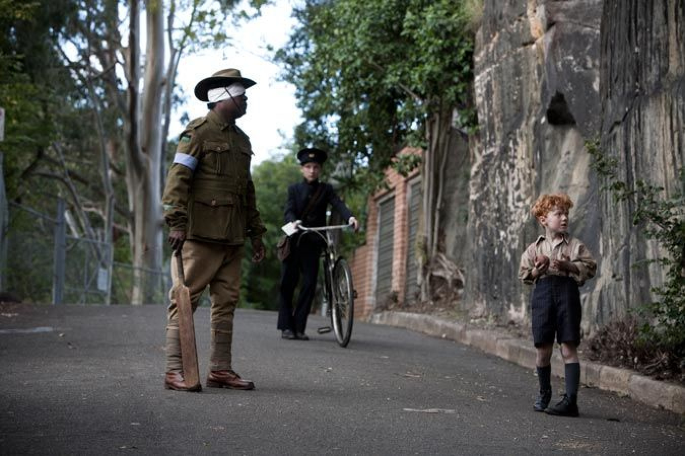 A soldier in military attire holds a cricket bat while standing on a quiet road, looking intently at a young boy dressed in vintage clothing and shorts. Behind them, a child in a dark outfit rides a bicycle, adding a sense of nostalgia to the scene, surrounded by greenery and brick walls.