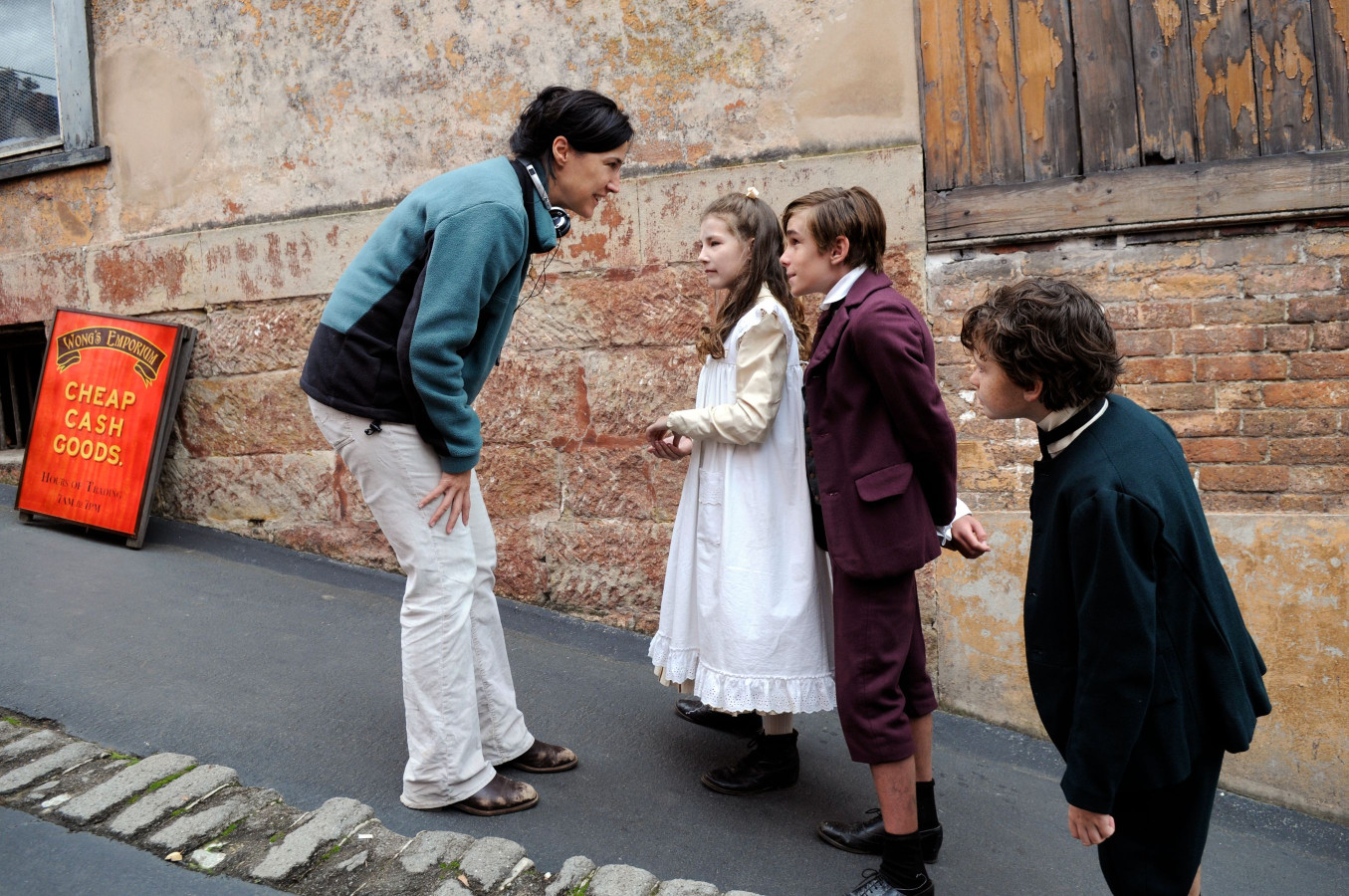 A woman interacts with three children in period costumes on a narrow street, surrounded by brick walls and a wooden door. A sign in the background advertises "Cheap Cash Goods." The woman appears to be giving direction or coaching the children, who are looking at her with expressions of curiosity.