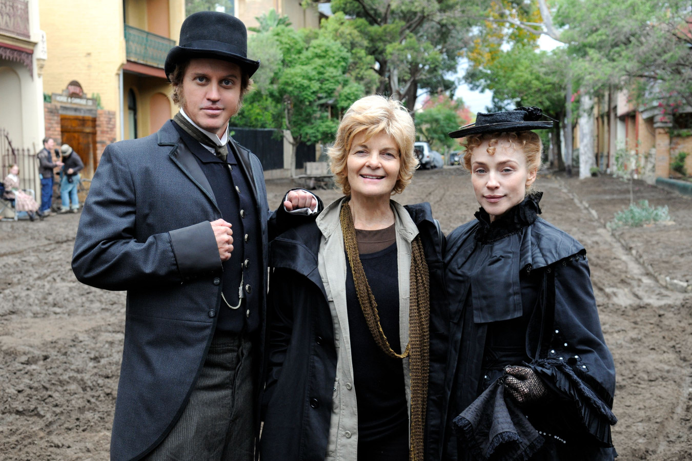 Three people pose together outdoors. The man on the left wears a vintage suit and a top hat, while the woman in the center has a warm smile and casual attire. The woman on the right is dressed in a black, vintage-style dress and hat. In the background, there are trees and a muddy path suggesting a film set environment.