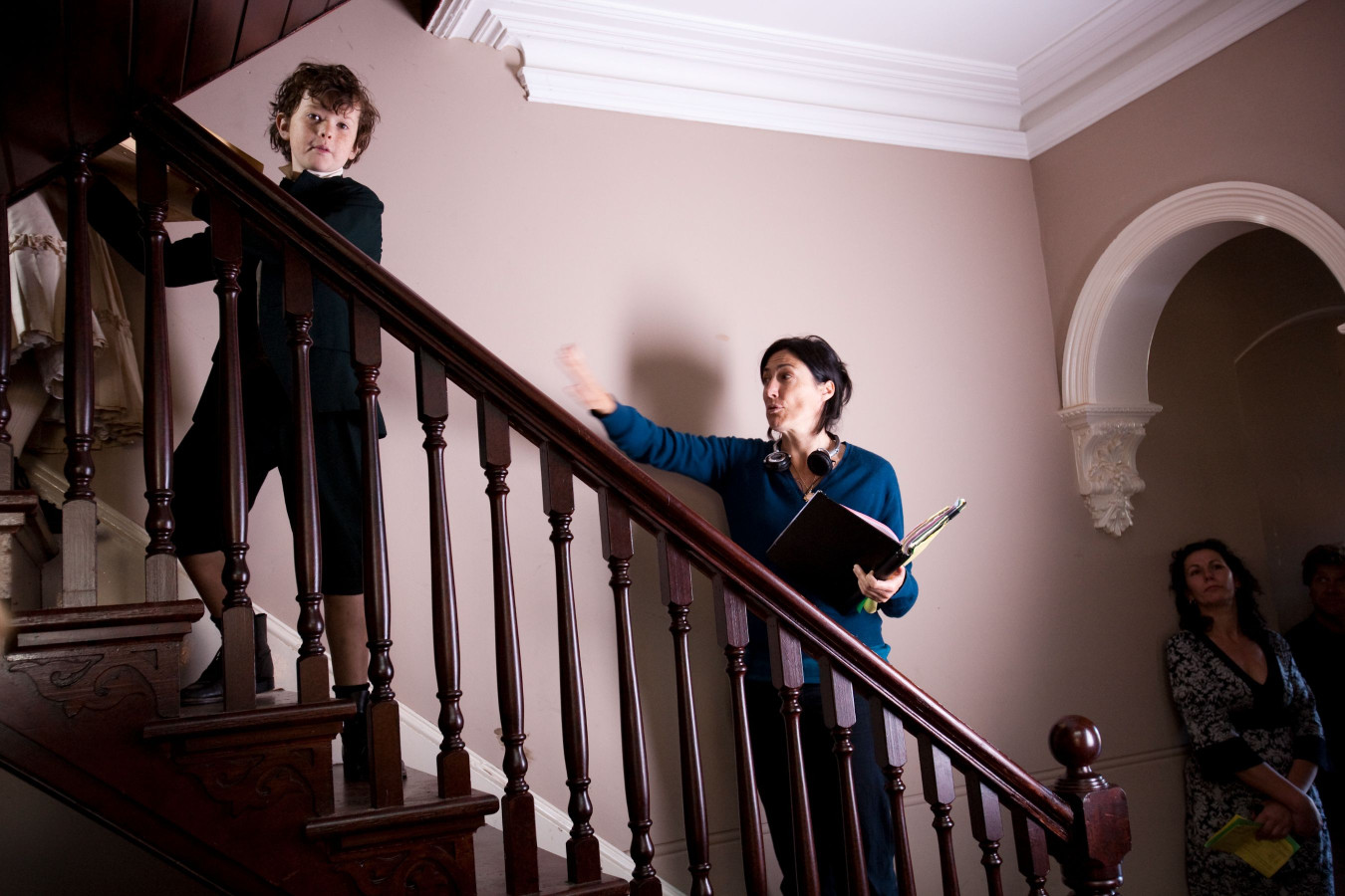 A young boy in a black outfit stands on the stairs, looking down at an adult woman at the bottom, who gestures while holding a binder. Behind her, two other individuals observe the interaction, creating a scene that appears to involve direction or discussion. The setting has warm tones and classic architectural details.