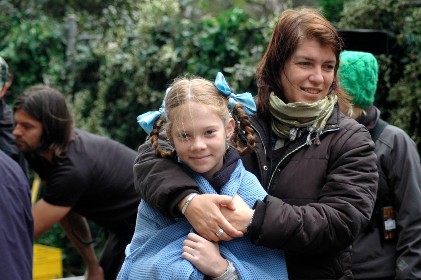 A girl with braids and blue bows smiles while wrapped in a blue blanket, standing close to a woman in a dark jacket and scarf, who embraces her from behind. In the background, blurred figures and greenery suggest an outdoor setting, likely on a film set.