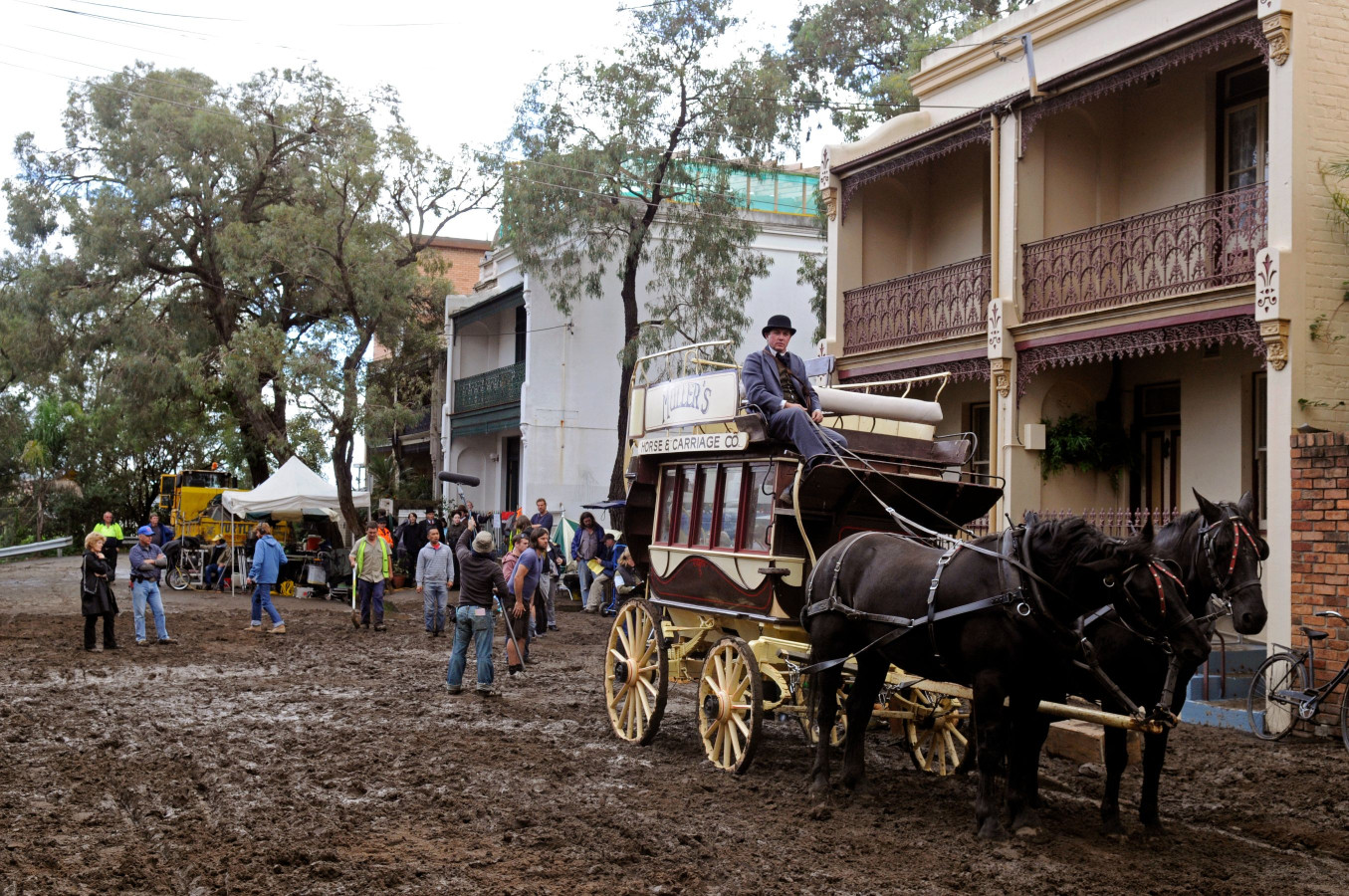 A horse-drawn carriage sits in a muddy street, with a driver wearing a hat on top. Surrounding the carriage are onlookers and tents, while historic buildings line the street under a cloudy sky.