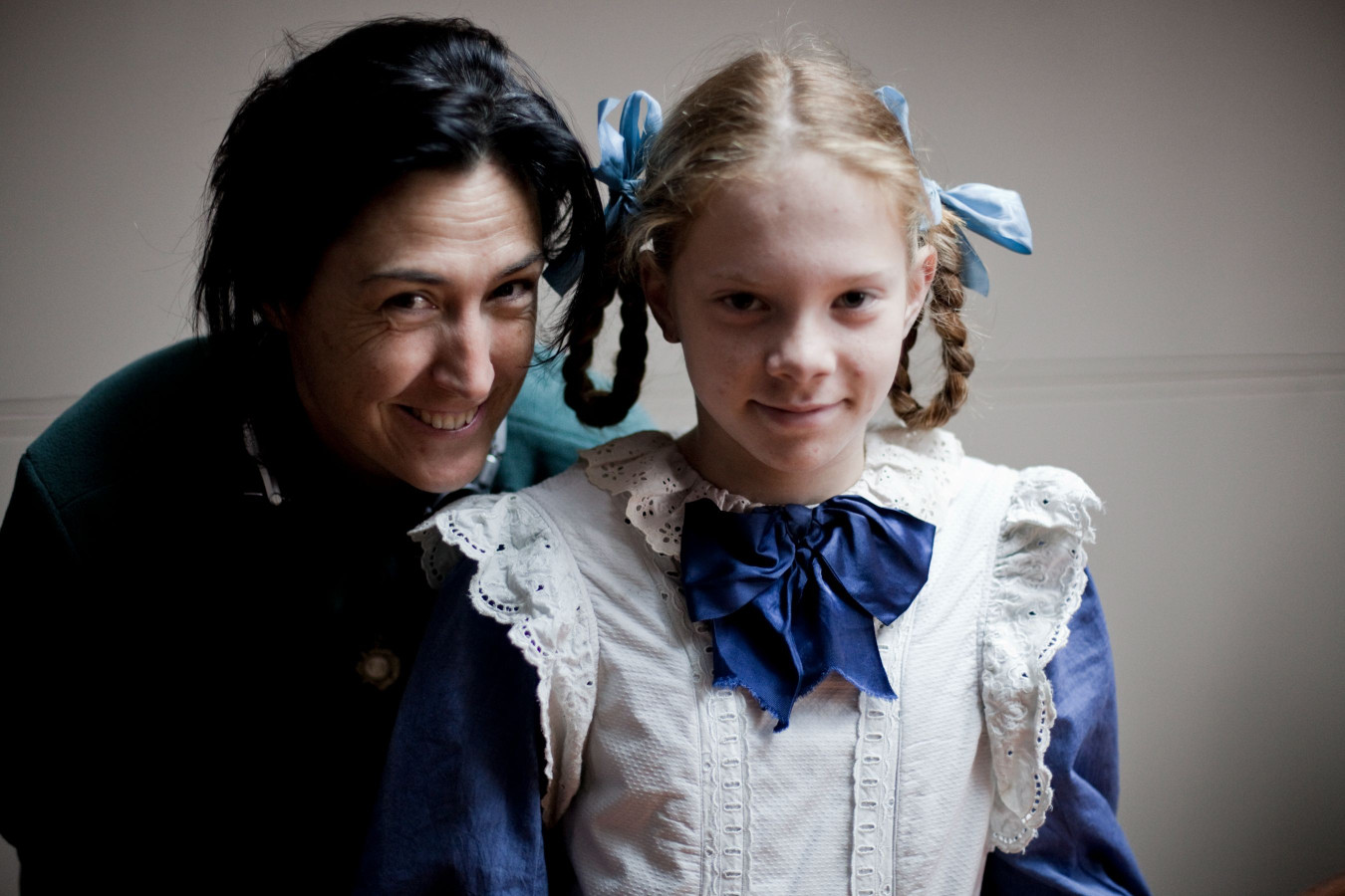 A woman with dark hair smiles joyfully while standing behind a young girl dressed in an old-fashioned outfit with pigtails tied with blue ribbons. The girl has a serious expression and wears a white dress with a large blue bow at the collar.