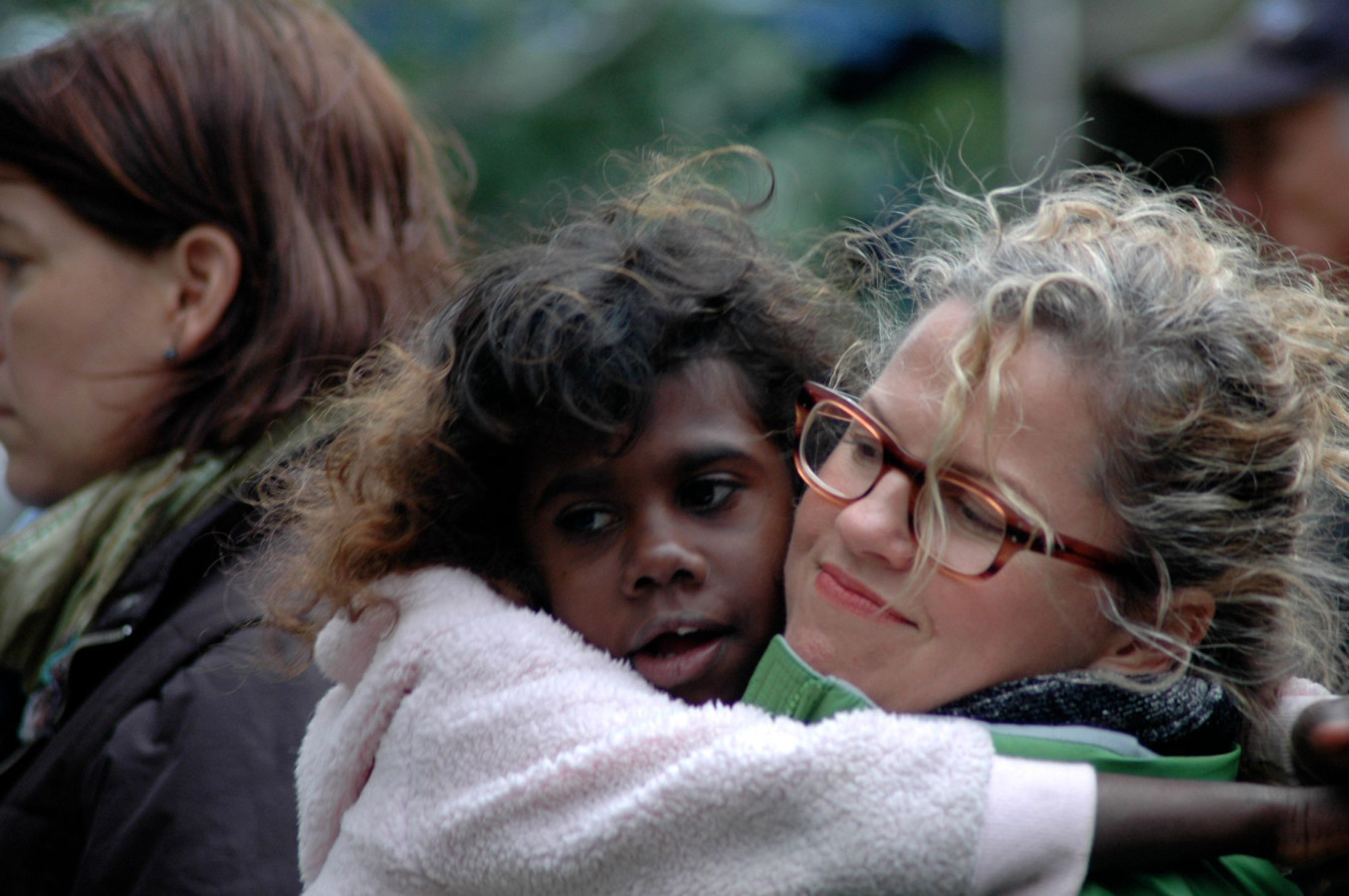 A young girl with curly hair is being embraced by a woman with curly, blonde hair and glasses, both smiling gently. In the background, there are blurred figures of other individuals, suggesting a gathering or event.