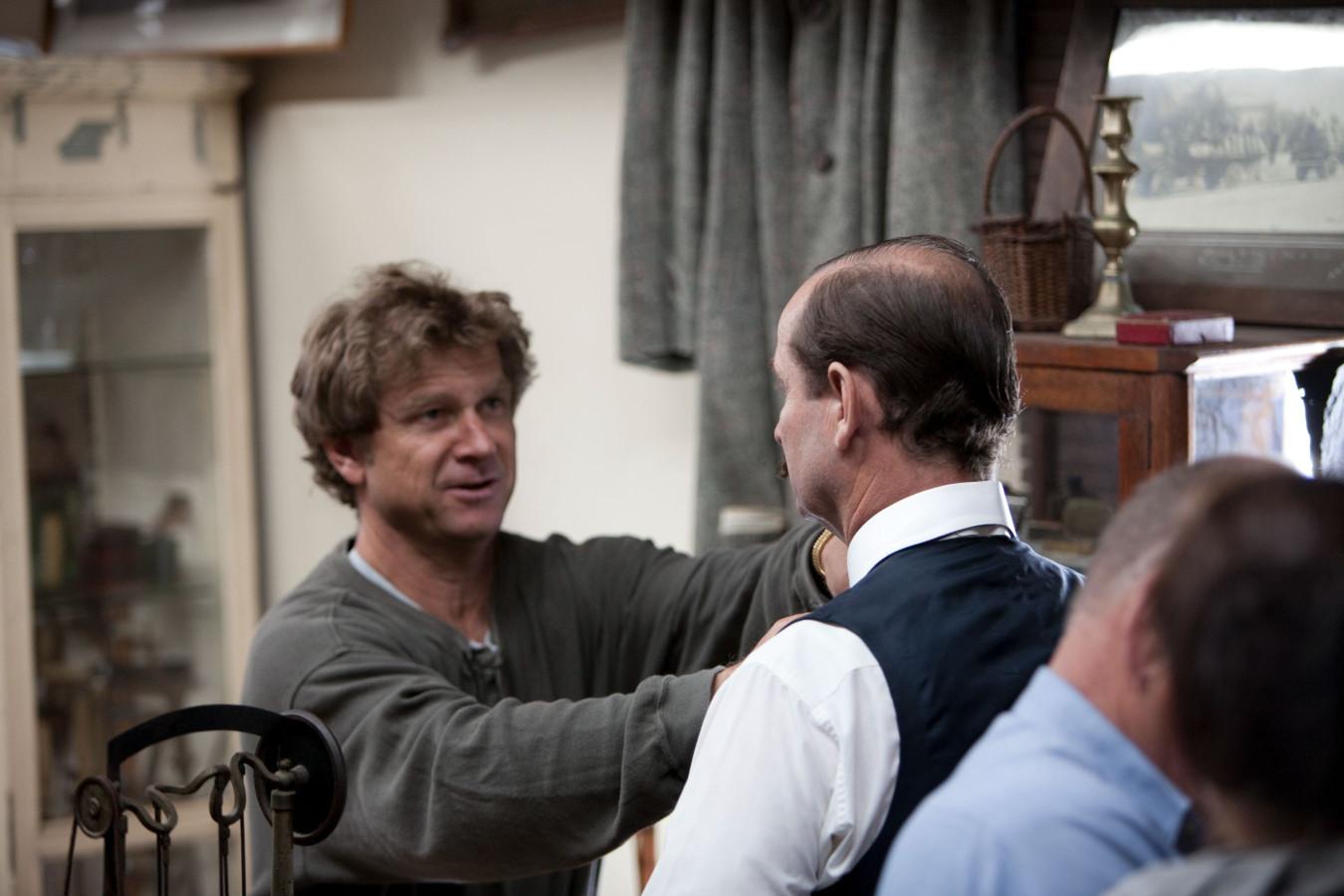 A man in a gray shirt adjusts the collar of another man wearing a formal vest and shirt. They are indoors, surrounded by vintage furnishings and several people in the background, engaged in a casual conversation.