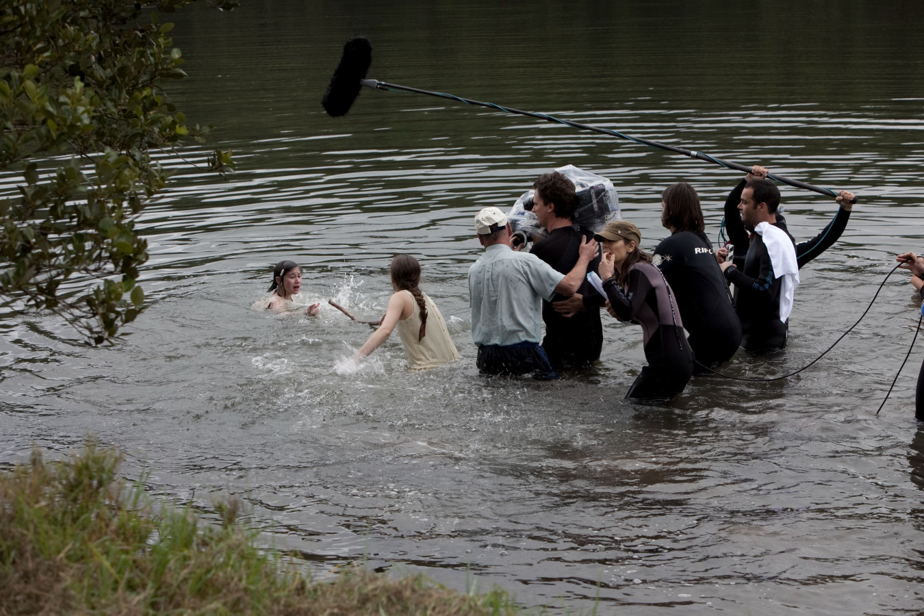 A group of people is gathered around a river, with two individuals partially submerged and appearing to act in a scene involving water. Crew members hold cameras and sound equipment, while others assist or observe the filming process. The setting is lush and natural, reflecting a day of shooting a scene in the outdoors.