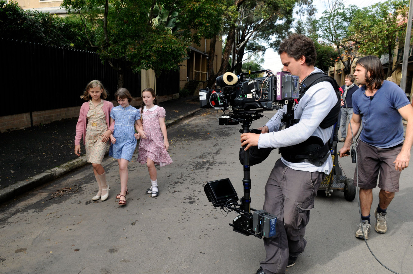 Three children dressed in vintage-style dresses walk together down a street. A cameraman equipped with a professional camera rig follows them closely, while another crew member assists in the background. Trees and buildings line the street, creating a lively filming atmosphere.