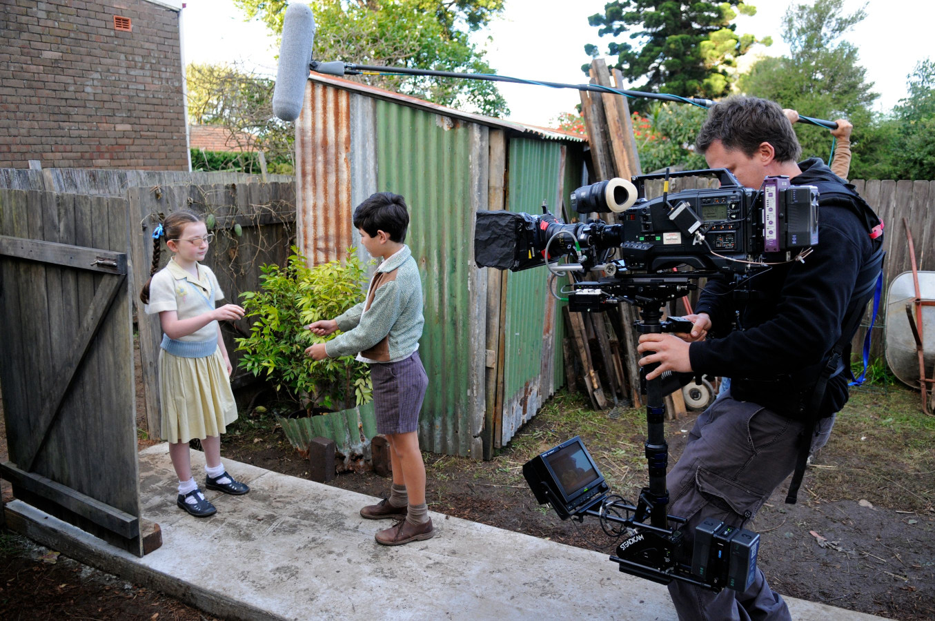 A young girl in glasses stands near a wooden gate, interacting with a boy who is holding out his hand, seemingly offering something. A film crew is positioned nearby, with a cameraman operating a large camera on a stabilizer, while a microphone is positioned overhead. The background features a rustic setting with corrugated metal structures and greenery.