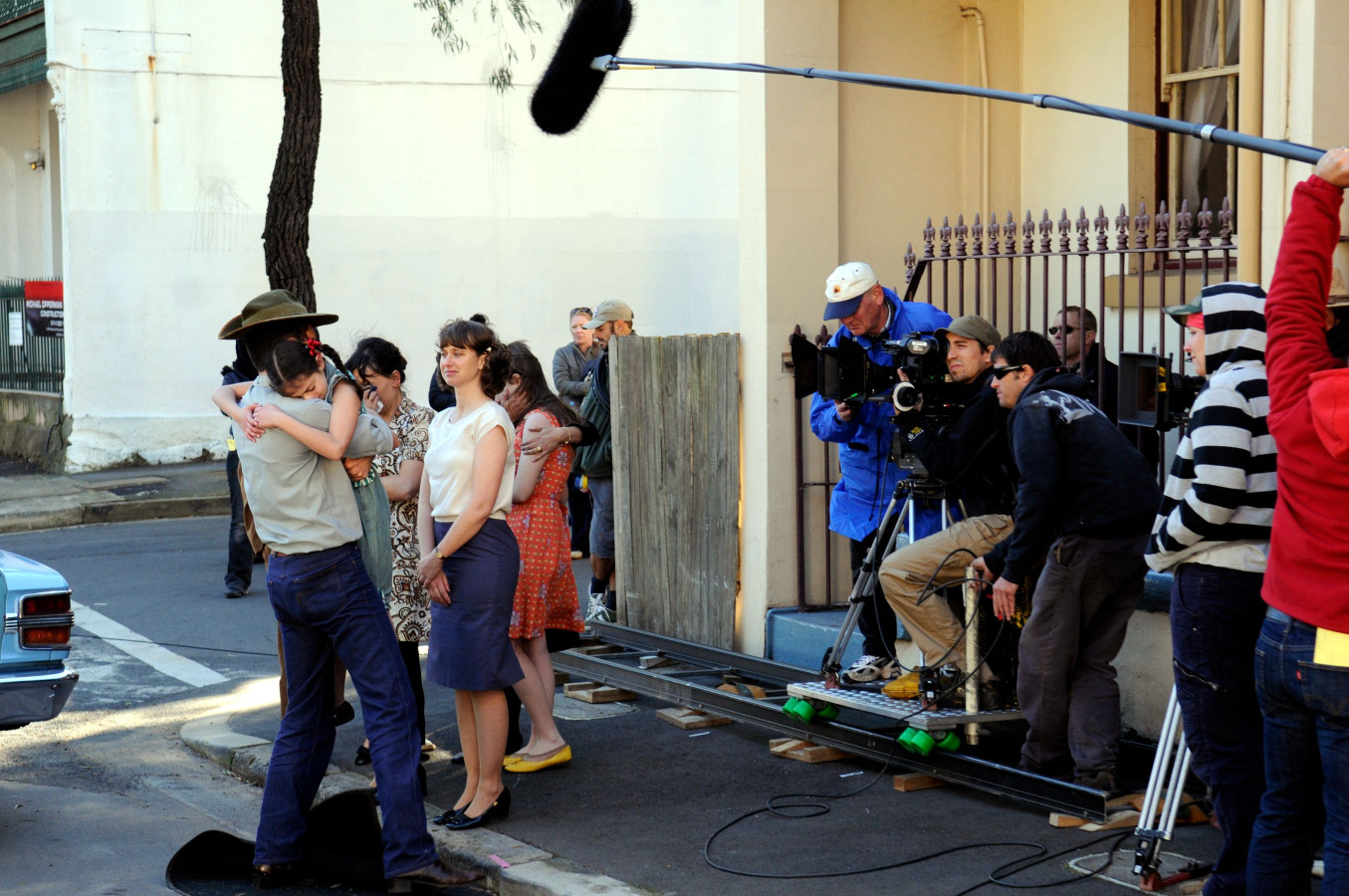 A film crew is on set, capturing a scene where a man in a cowboy hat embraces a woman, while several other women stand nearby, ranging in expressions from emotional to serious. Behind them, crew members manage cameras and sound equipment, contributing to the production process. The setting features a street corner with a blue car and a building facade in the background.