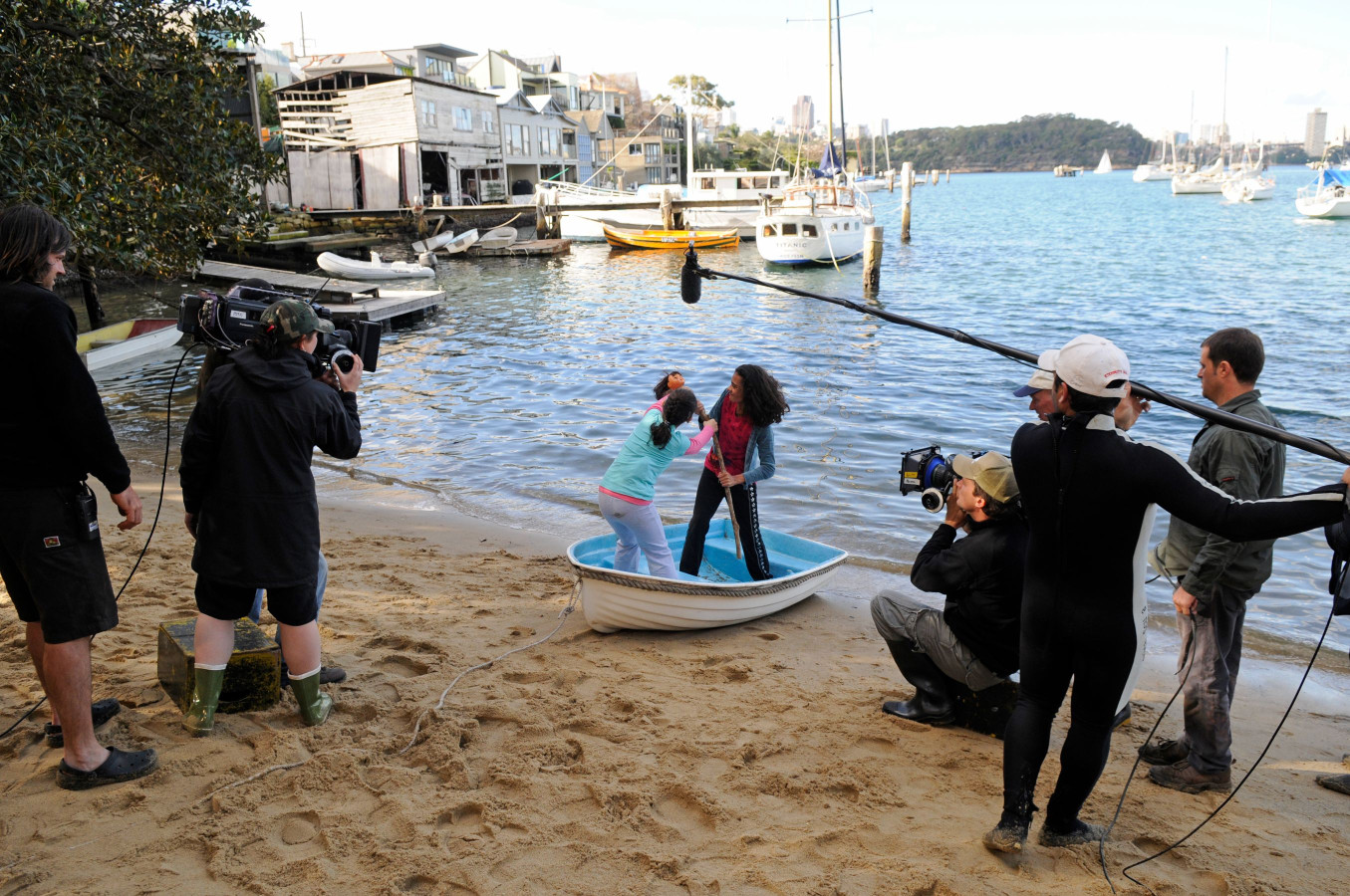 Two girls are playfully interacting in a small boat near the shore, surrounded by a sandy beach. A film crew is capturing the scene, with crew members operating cameras and holding microphones, while several boats are anchored in the background against a scenic waterfront.