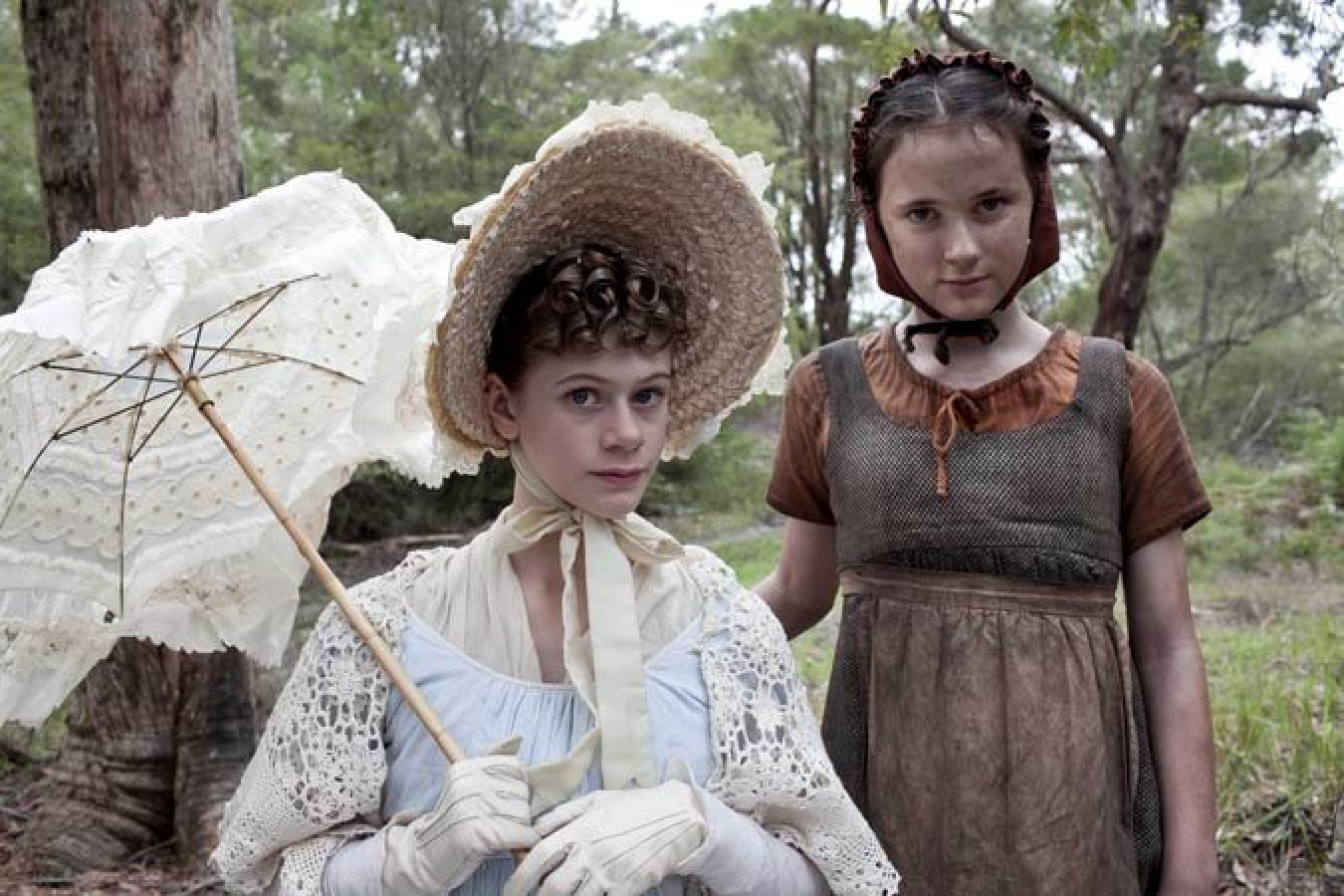 Two young girls stand in a forested area, one wearing a light blue dress and a white lace shawl, holding a delicate lace parasol, while the other is dressed in a simpler brown outfit with a fitted bodice and a red bonnet. The scene conveys a historical vibe, set against a backdrop of greenery.