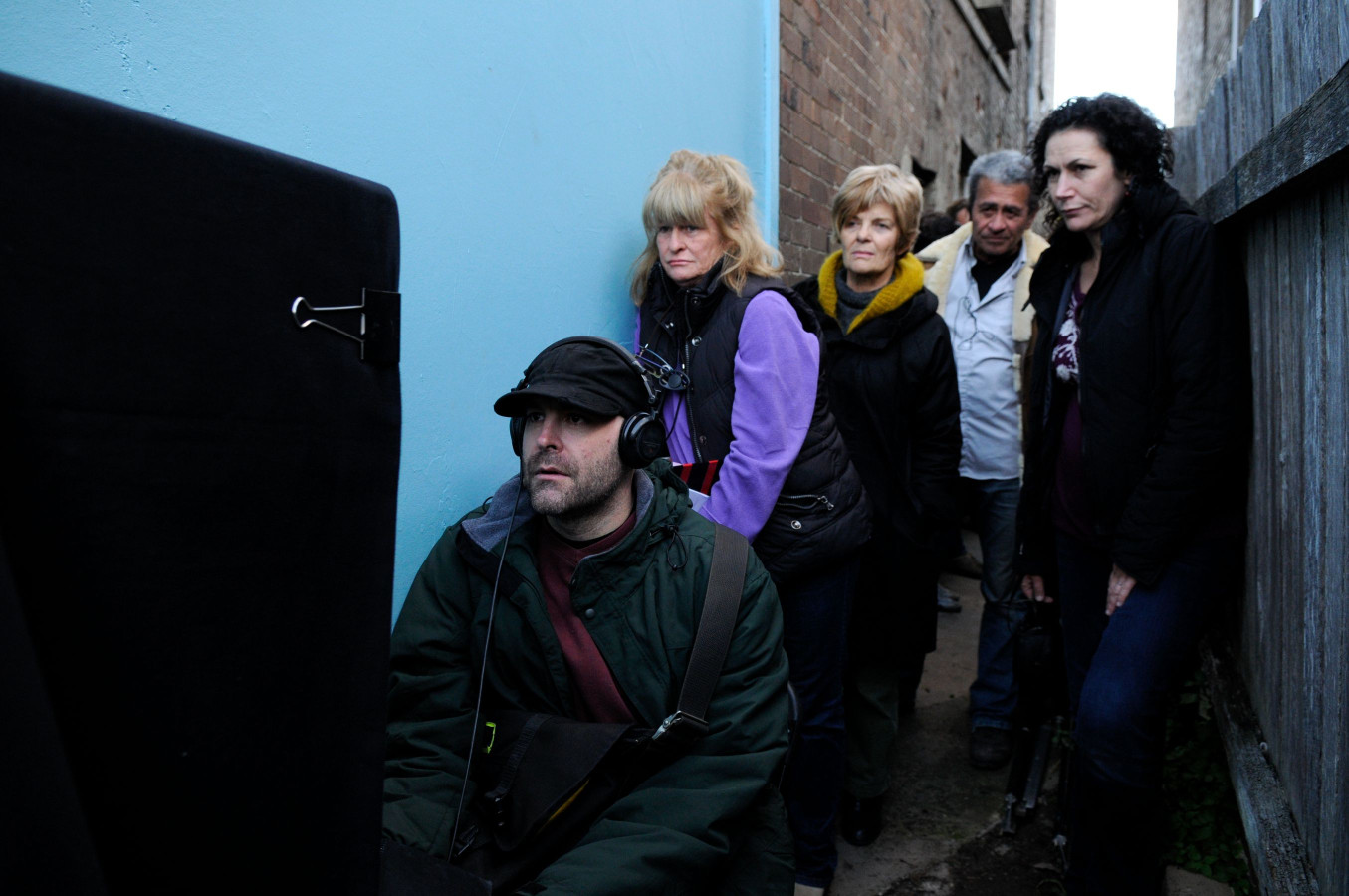 A group of five people watches intently as a man seated below them operates a camera or monitor in a narrow alley. The group includes three women and two men, with a mix of expressions ranging from curiosity to concern, while the scene is framed by a backdrop of brick walls and a light blue surface.