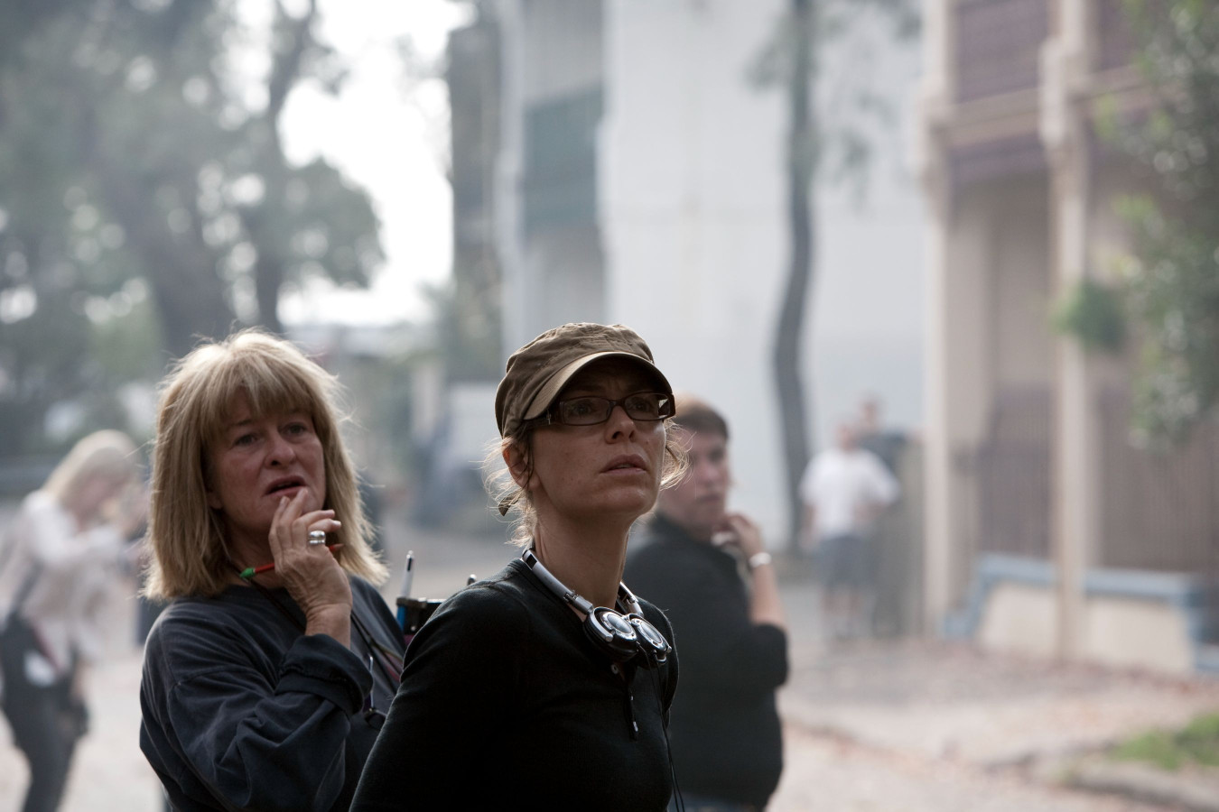 A group of people stand outdoors, some looking up with expressions of concern. One woman in the foreground, wearing a cap and glasses, appears focused and contemplative, while another woman nearby seems to be deep in thought, holding a pen. The background is hazy, suggesting a smoky atmosphere.
