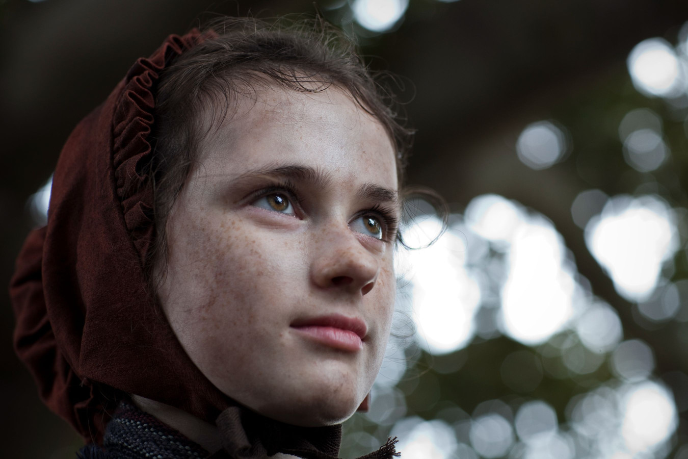 A young girl with curly hair peeks out from under a brown bonnet, her freckled face illuminated with a soft, thoughtful expression. The background features blurred greenery, creating a serene atmosphere.