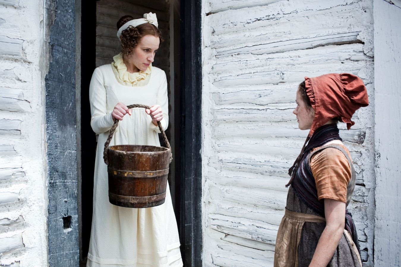 A woman in a long, white dress holds a wooden bucket while standing in a doorway, looking down at a girl with a red bonnet and a brown dress who stands outside. The background features a rustic, weathered wooden wall.