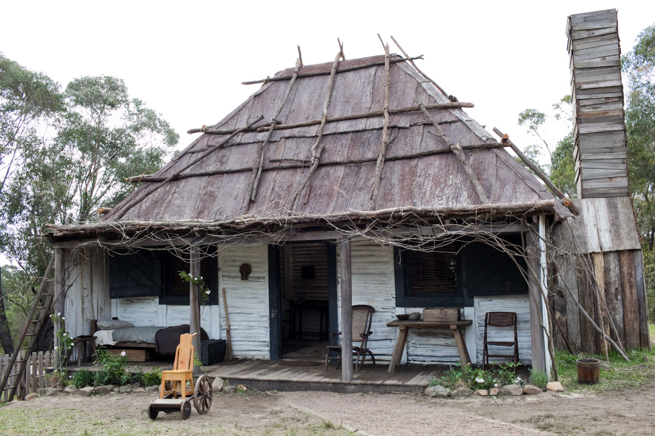A rustic wooden cottage features a thatched roof made from branches and straw. The front porch has a small table and chairs, with a cart in the foreground and surrounding greenery. A tall, narrow chimney rises from the side of the house, blending into a serene natural setting.