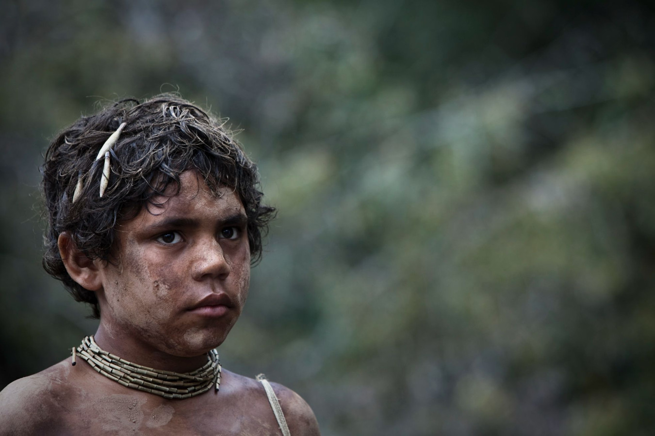 A young boy with curly, unkempt hair and dirt smeared across his face stands in a natural setting. He wears handmade accessories around his neck and has twigs or small sticks in his hair, gazing intently into the distance.
