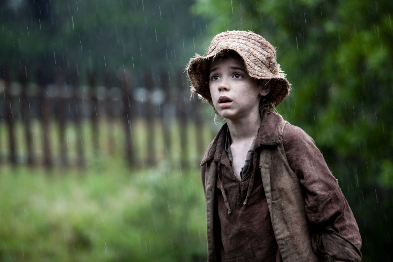 A young boy with a straw hat stands in the rain, looking up with a concerned expression. He wears a worn, dirty shirt and vest, and the blurred background features a grassy area and a wooden fence, emphasizing the somber mood of the scene.