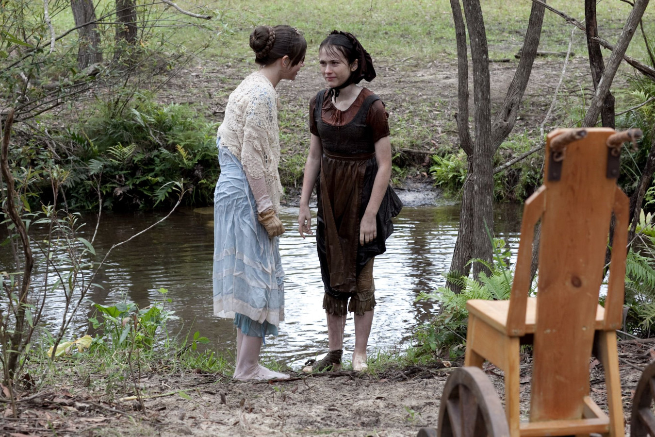 Two young girls stand beside a shallow stream in a wooded area. One girl, dressed in a light blue dress, appears to be speaking intensely to the other, who is wearing a dark, wet dress. Their expressions suggest a moment of confrontation or serious discussion, with a wooden chair and other elements of the natural setting visible nearby.