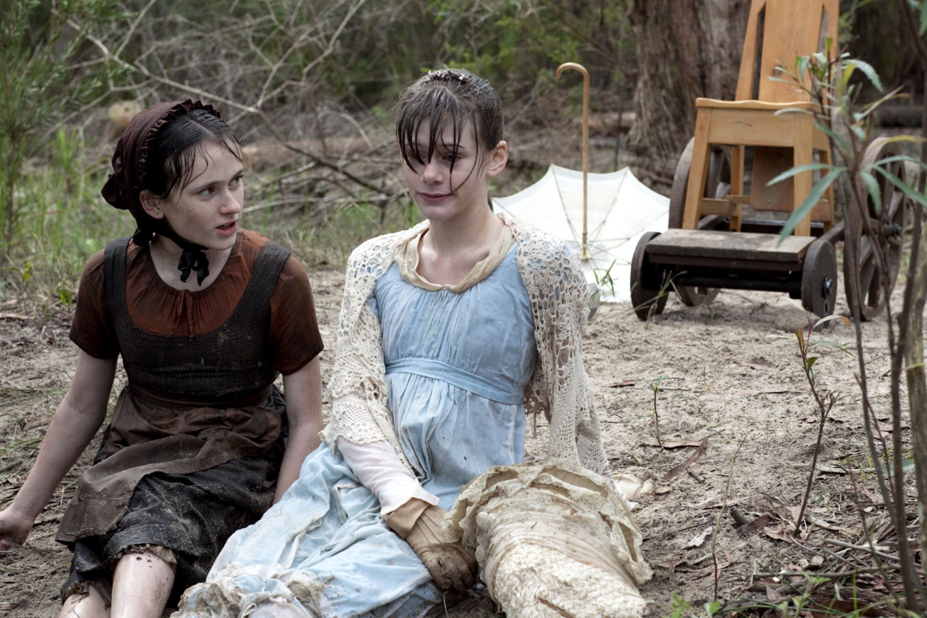 Two girls, one in a brown dress and the other in a light blue gown, sit on the ground surrounded by trees. The girl in brown looks attentively at the girl in blue, who appears distressed and has wet hair, while an abandoned wooden chair and a partially visible umbrella are nearby.