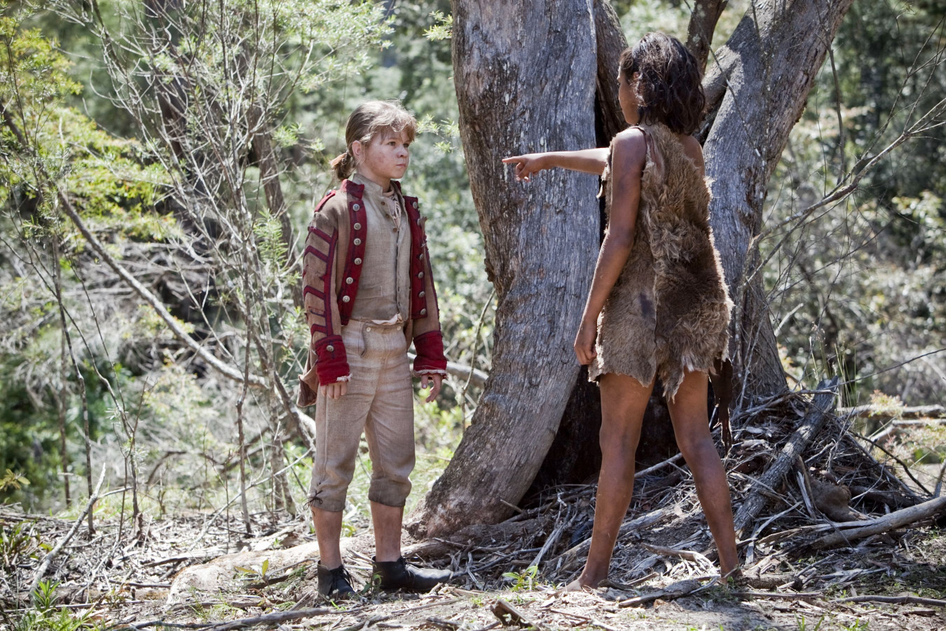A young boy in a military-style jacket stands facing a girl dressed in ragged clothing made from animal hide. The girl points assertively at him, while both are positioned near a large tree in a forested area.