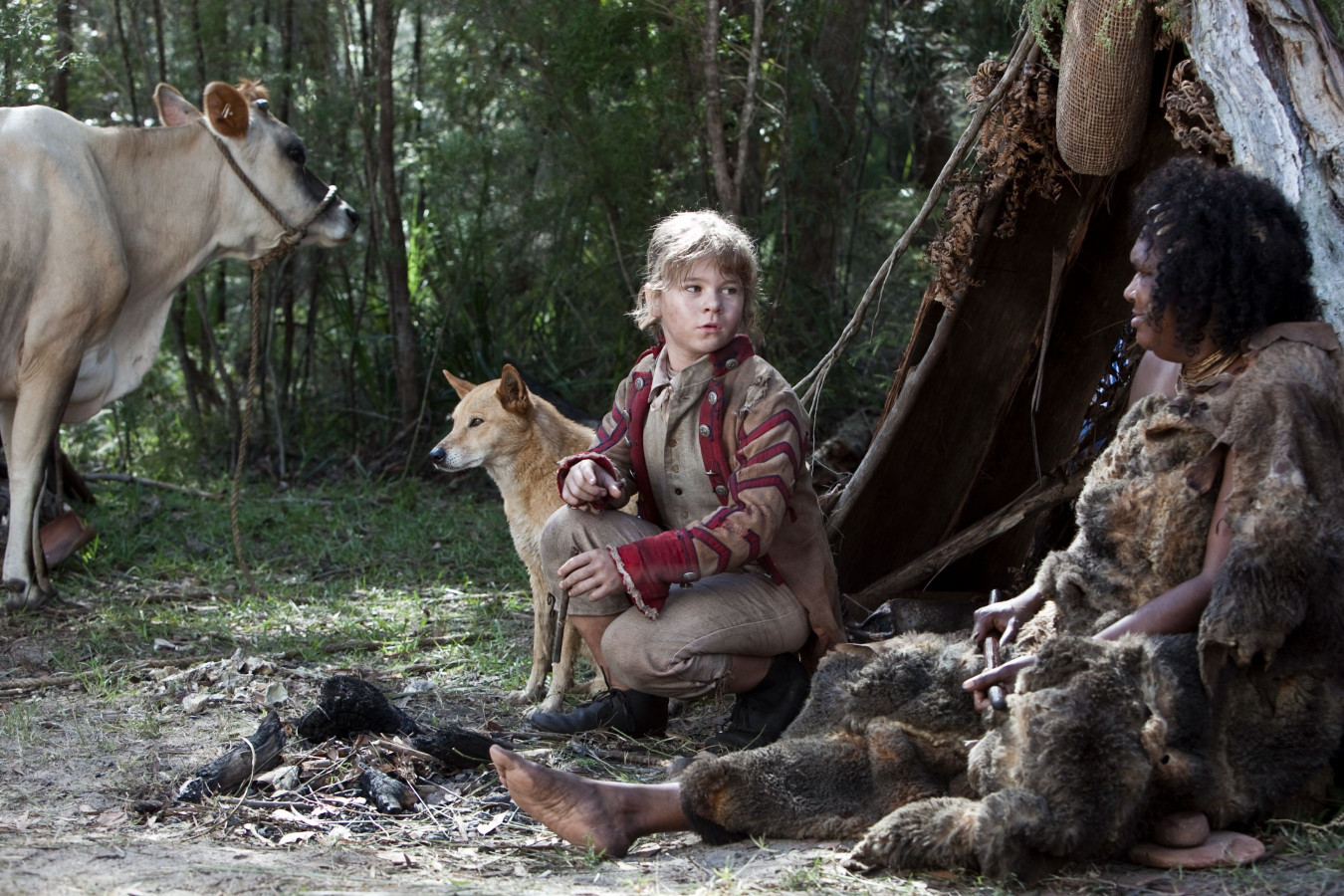 A young child sits on the ground beside a dingo, looking intently towards a nearby cow. An adult person is seated nearby, partially covered in fur, with a fire pit visible between them in a forested setting.