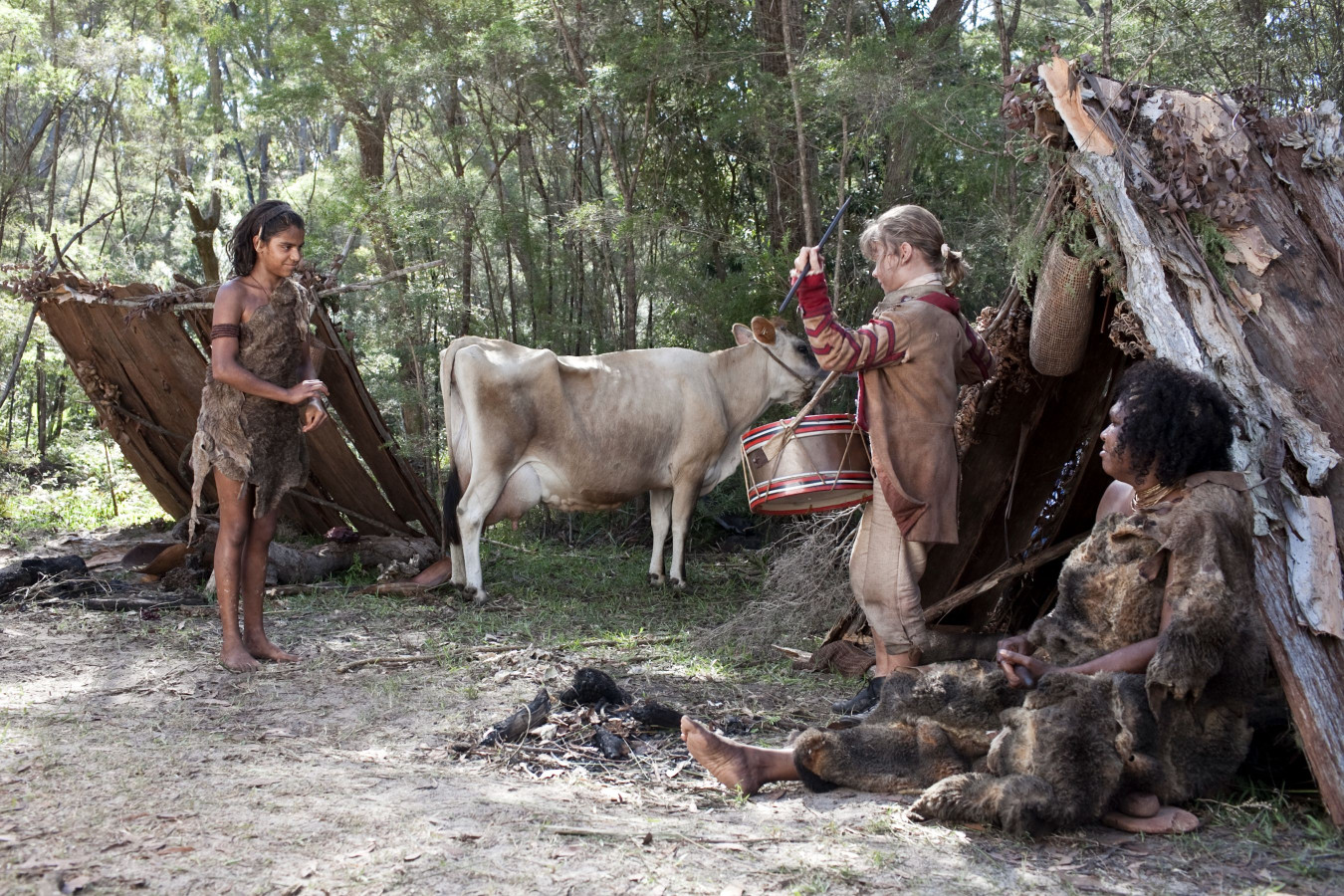 A young boy in primitive attire stands near a cow, while a girl in similar clothing plays a drum and appears to be engaging with the cow. Two other individuals, one sitting and another partially hidden, are near makeshift shelters made of branches and leaves in a forested area.