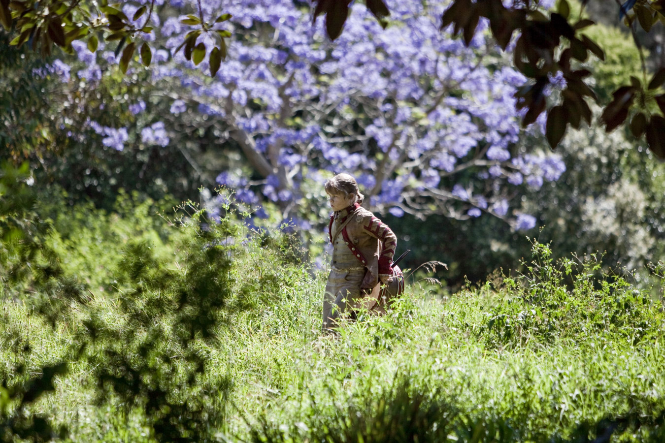 A child walks through a lush green field, surrounded by tall grass and vibrant foliage. In the background, a tree is adorned with bright purple flowers, creating a picturesque scene of nature.