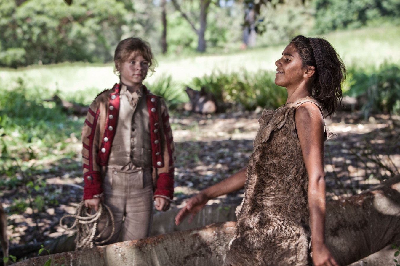 A young boy in a red and beige military-style jacket stands in a forest clearing, looking at a girl dressed in a tattered, brown garment. The girl, with her hair down and a joyful expression, stands beside a fallen log, surrounded by greenery and sunlight filtering through the trees.