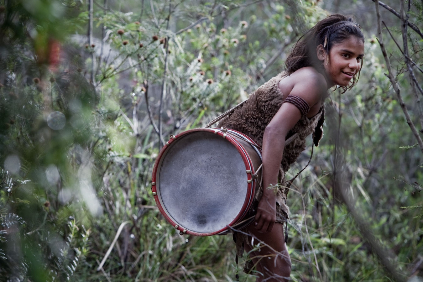 A young girl stands in a dense, green forest, wearing a simple, earth-toned outfit. She is smiling and holding a large drum, positioning it as if ready to play. The surrounding vegetation includes tall grasses and small shrubs, creating a lively natural backdrop.