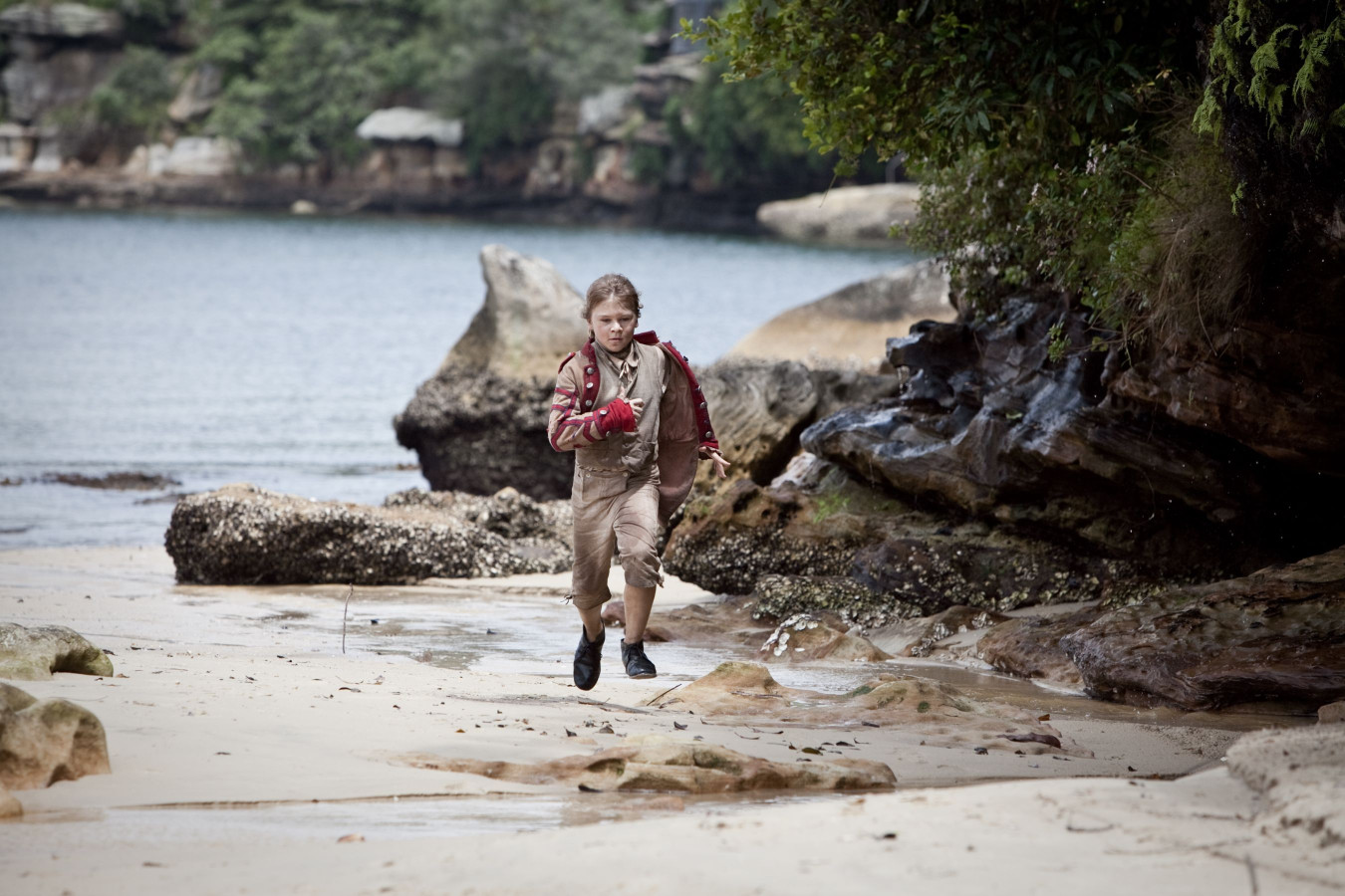 A young boy in a brown shirt and shorts runs along a sandy beach, surrounded by rocky formations and shallow water. Lush greenery is visible in the background, and the scene conveys a sense of adventure and playfulness.