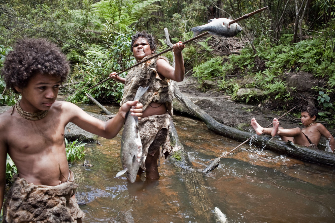 A group of children is wading in a shallow stream, engaged in fishing. One child holds a spear with a fish, while another poses triumphantly with a catch. Surrounding vegetation includes ferns and trees, creating a natural, lush backdrop.