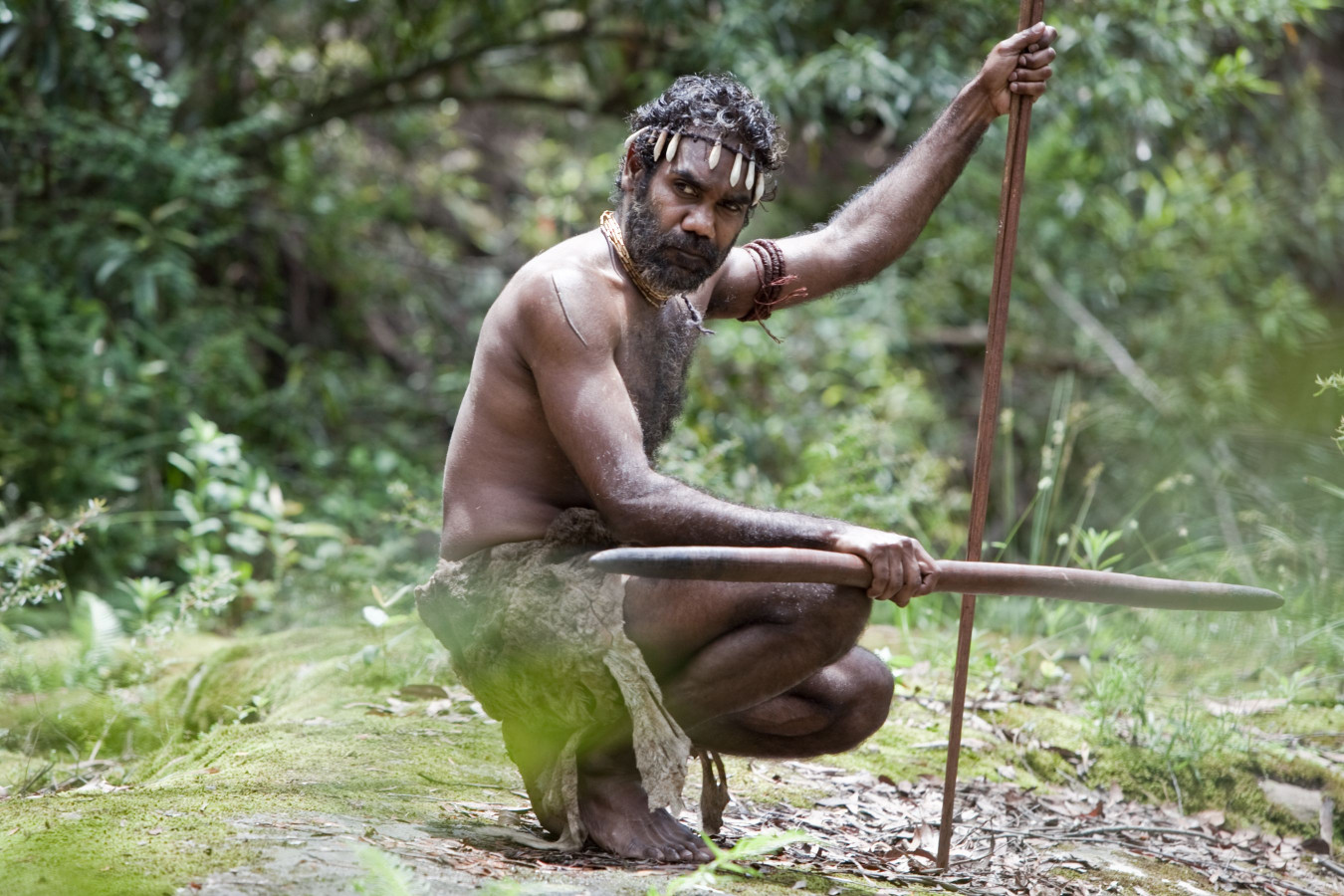 A man dressed in traditional garb sits in a forest, holding a spear while crouching on the ground. His hair is adorned with ornaments, and he appears focused and alert, surrounded by greenery and natural elements.
