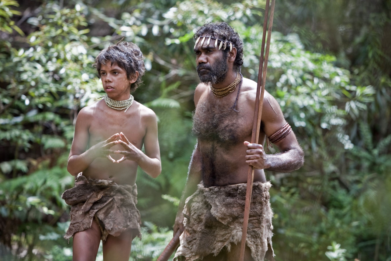 A young boy and a man, both wearing simple, textured attire made from natural materials, stand among lush greenery. The boy appears contemplative, gesturing with his hands, while the man holds a spear and looks attentively into the distance.