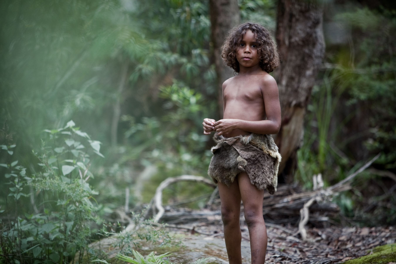 A young child stands in a lush, green forest, wearing a simple garment made from animal hide. The child has curly hair and appears to be observing the surroundings with a curious expression.