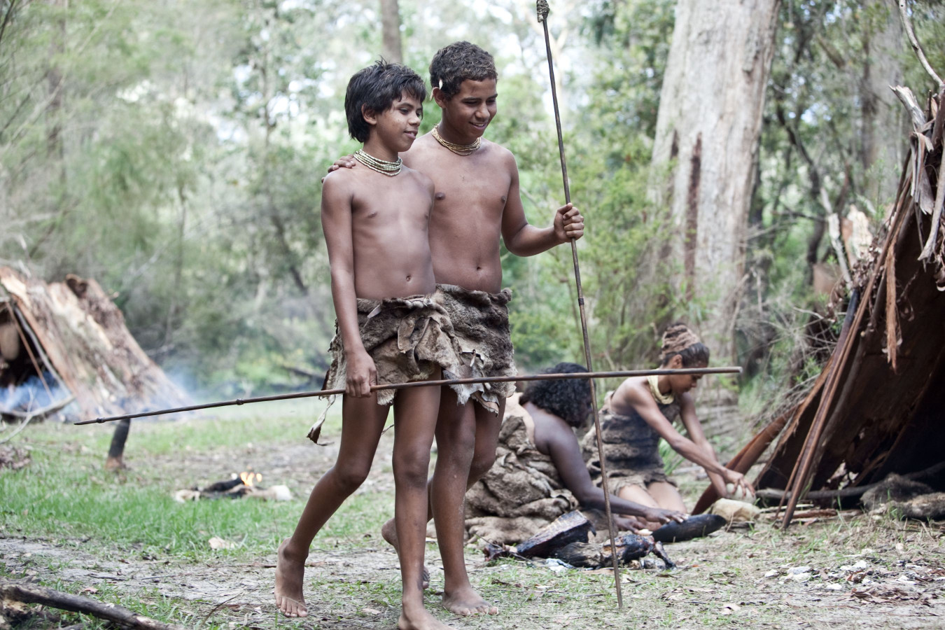 Two young boys stand together in a natural setting, wearing primitive clothing made of animal hides. One boy holds a spear, and they both appear to be smiling. In the background, another person is engaged in preparing food near a small fire, while a shelter made of branches is visible nearby.