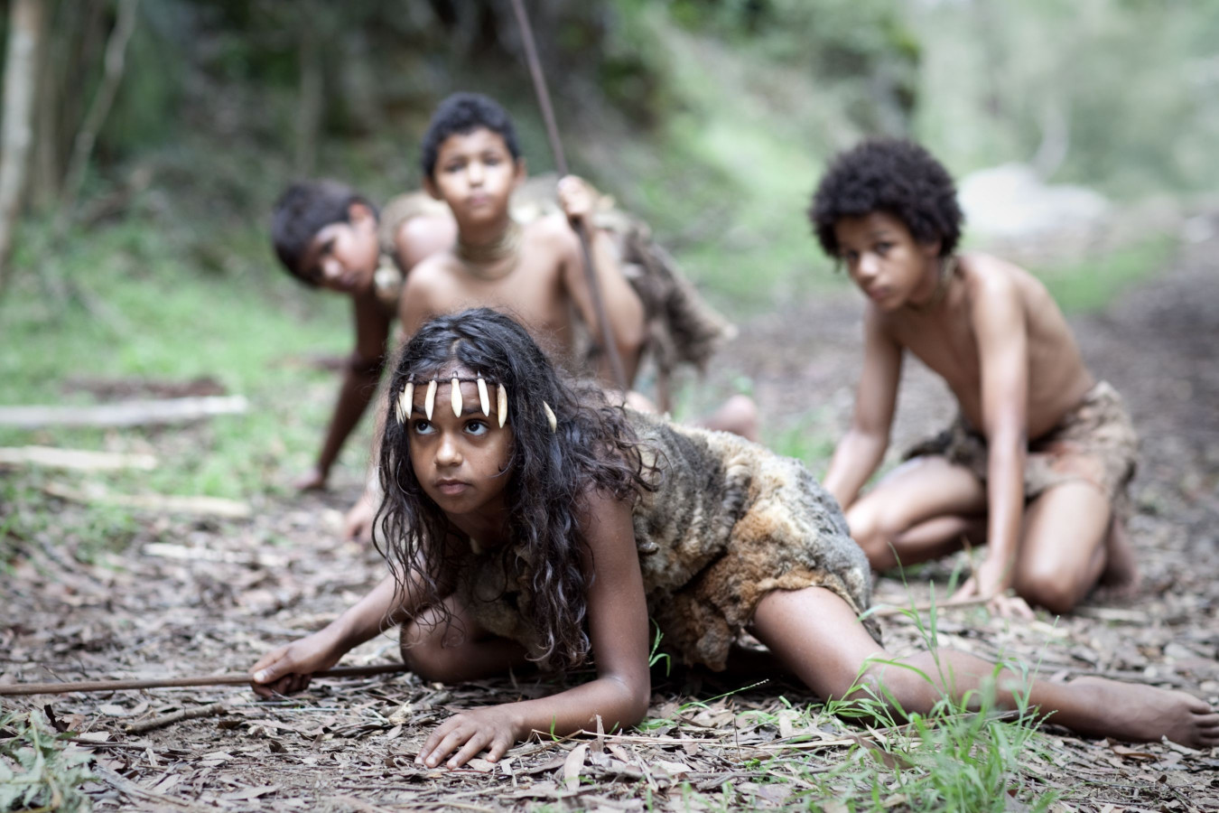 Four children are depicted in a lush forest setting, dressed in simple, animal skin clothing. They appear to be engaged in a playful activity, with one child on the ground holding a stick, while others are positioned in a crouched stance, observing curiously. The atmosphere captures a sense of exploration and adventure.