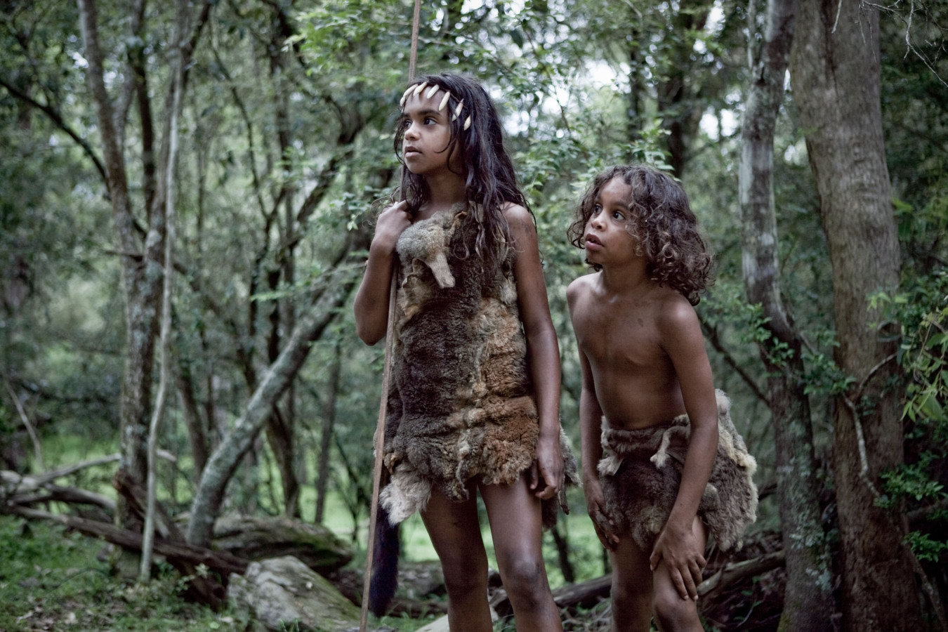 Two young children are dressed in primitive clothing made from animal skins, standing in a dense forest surrounded by trees. One child holds a spear and looks attentively at something in the distance, while the other appears curious, observing their surroundings.