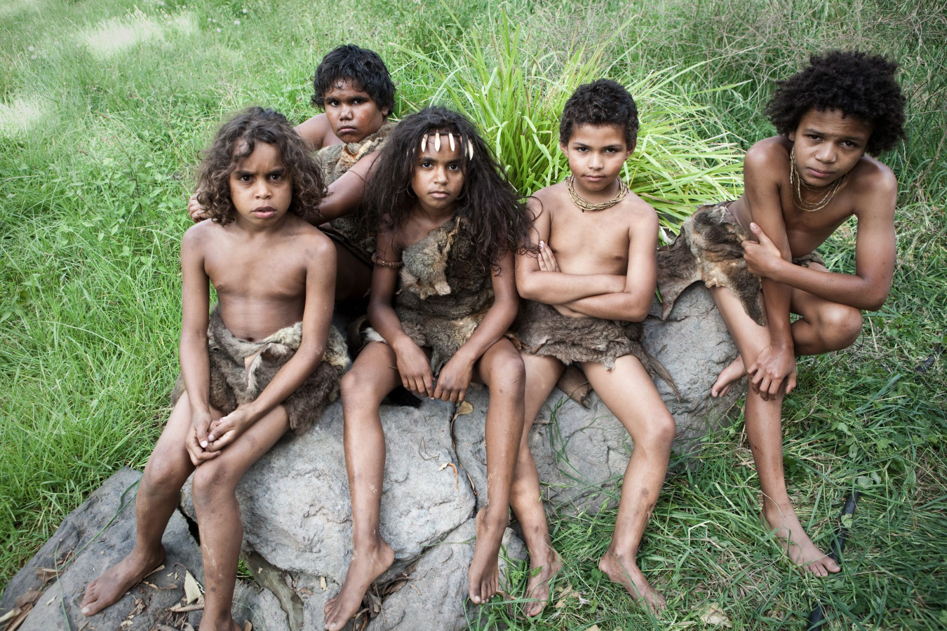 Five children sit on a large rock in a grassy area, wearing minimal clothing made of animal skins. They exhibit serious expressions, with some adorned with simple accessories, and display a range of hair textures. The background features lush greenery.