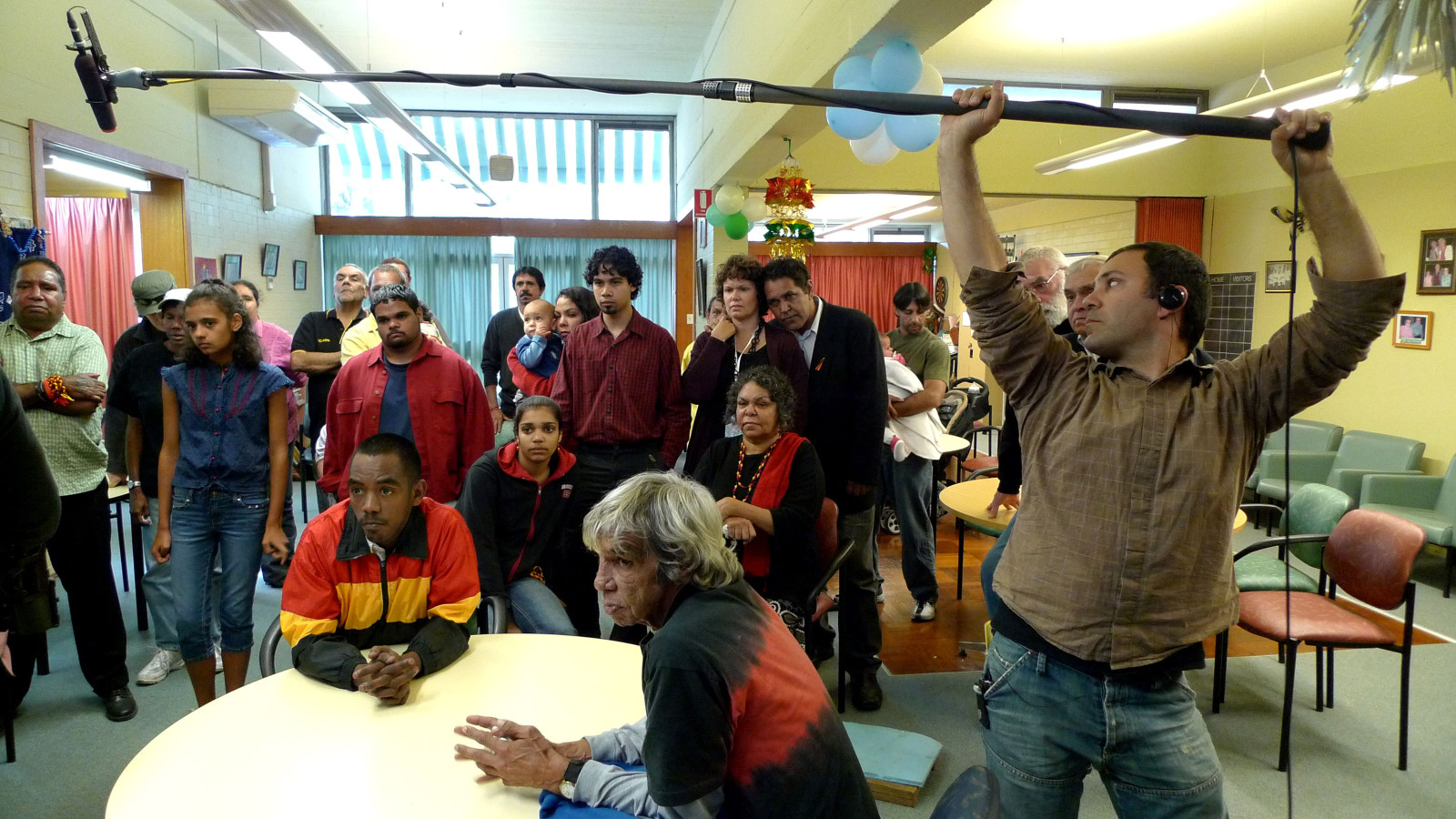 A diverse group of people is gathered in a room, with some observing intently while others sit at a table. A sound technician is holding a microphone boom overhead, capturing the scene. The atmosphere suggests a film shoot or a community event, with decorations visible in the background.