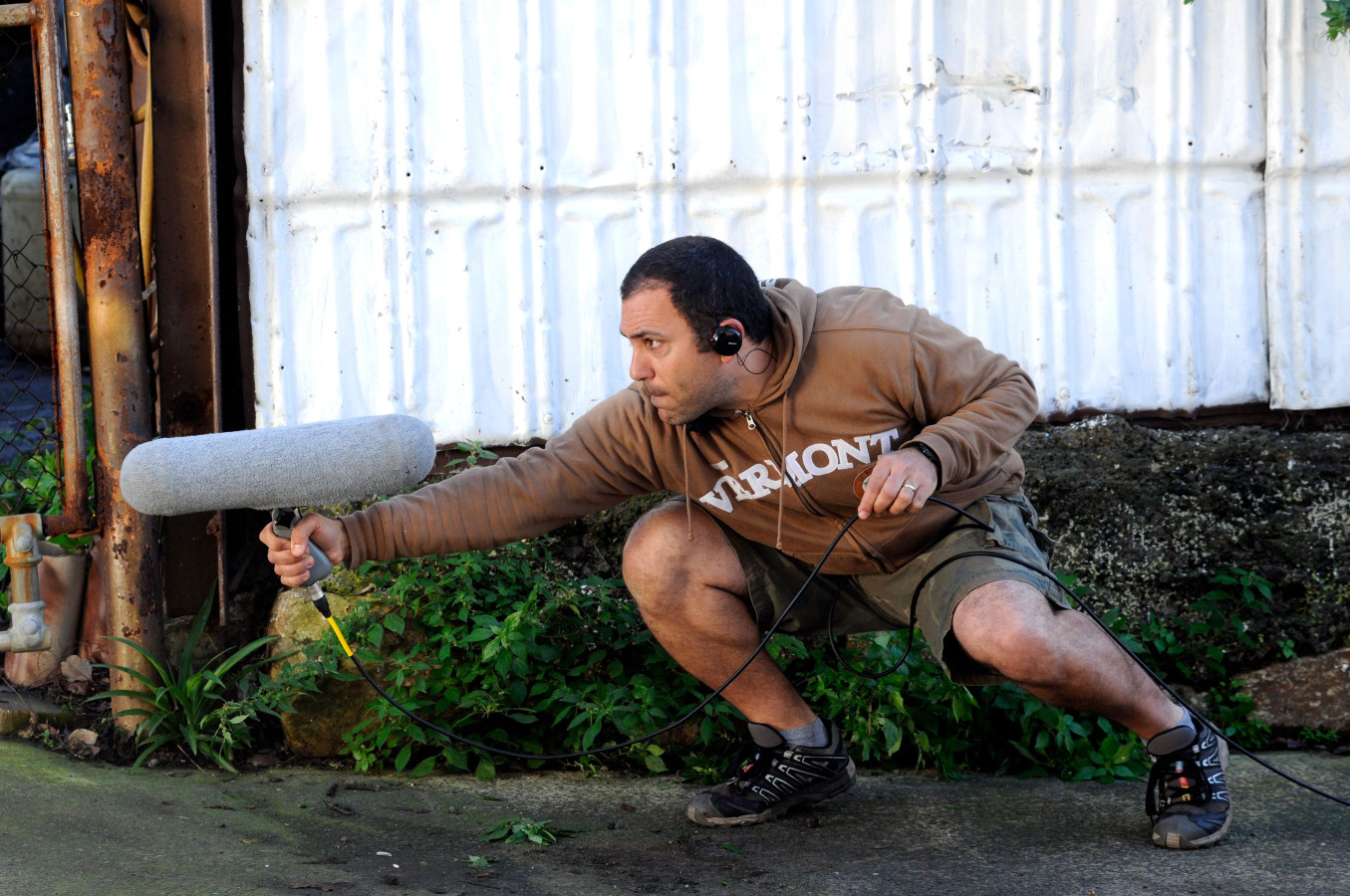 A man in a brown hoodie squats low to the ground, holding a microphone on a boom pole while wearing headphones. He appears to be focused and attentive, with lush greenery and a corrugated metal backdrop behind him.