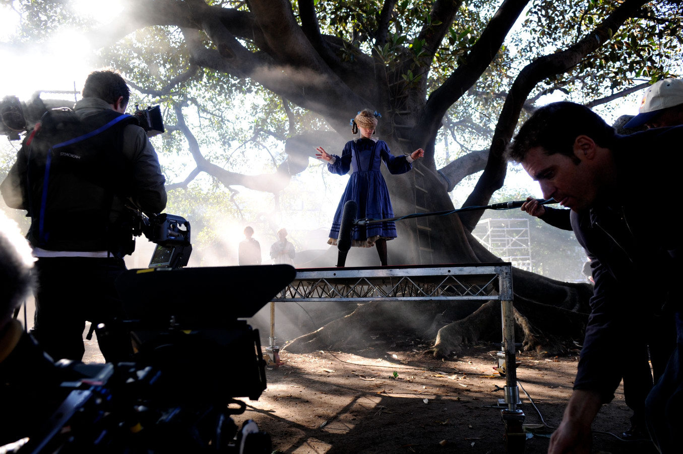 A child in a blue dress stands on a platform, performing theatrically under a large tree. Surrounding her, crew members operate cameras and equipment, with mist swirling in the background, suggesting an outdoor film or theater production.